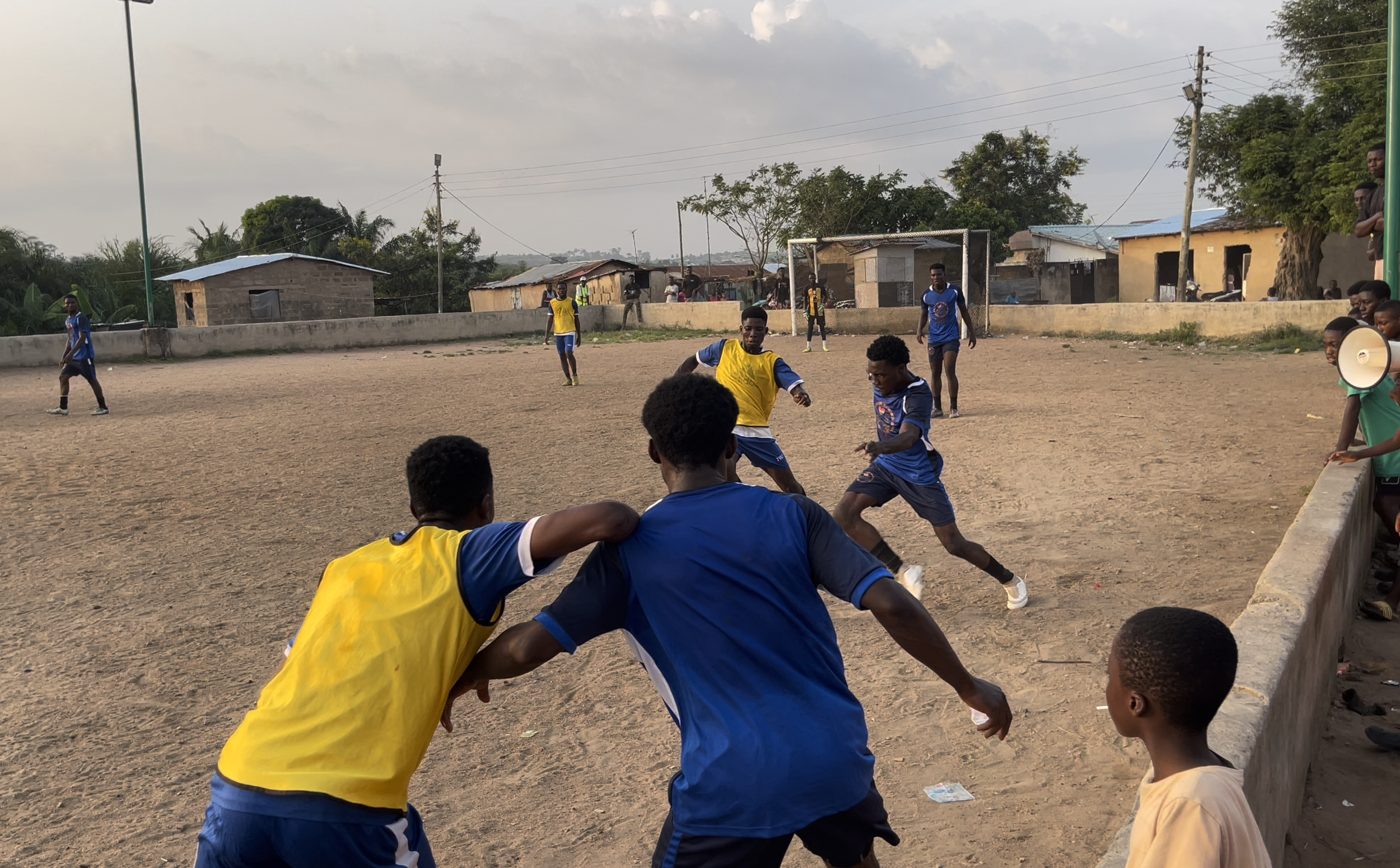 Young men playing soccer on a dirt field with some spectators watching from the sidelines.