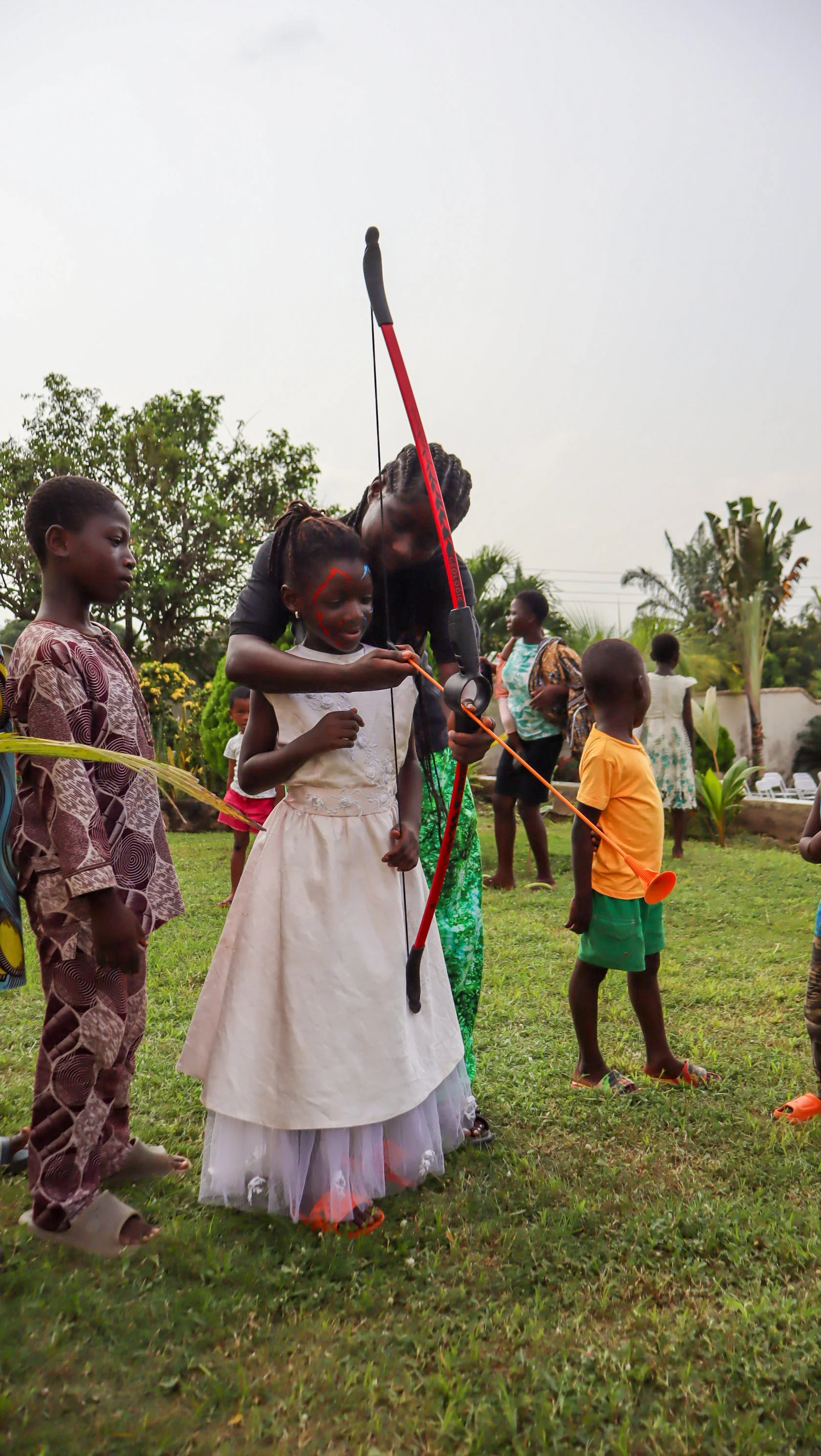 A woman teaching children how to shoot a bow and arrow outdoors in a grassy area, with several children and adults watching.