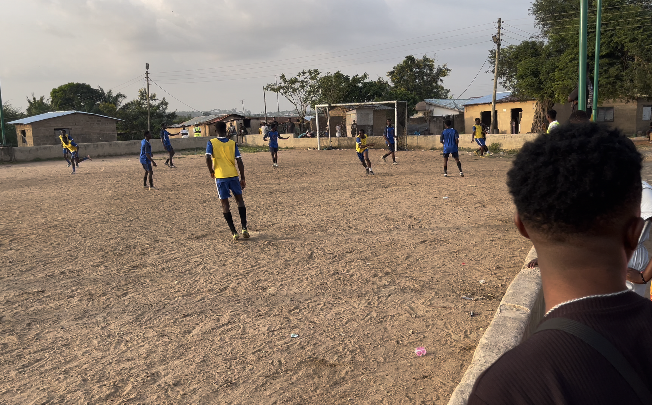 Youth soccer game on a dirt field with players in blue and yellow uniforms, some chasing the ball, others watching, near a small village with houses and trees.