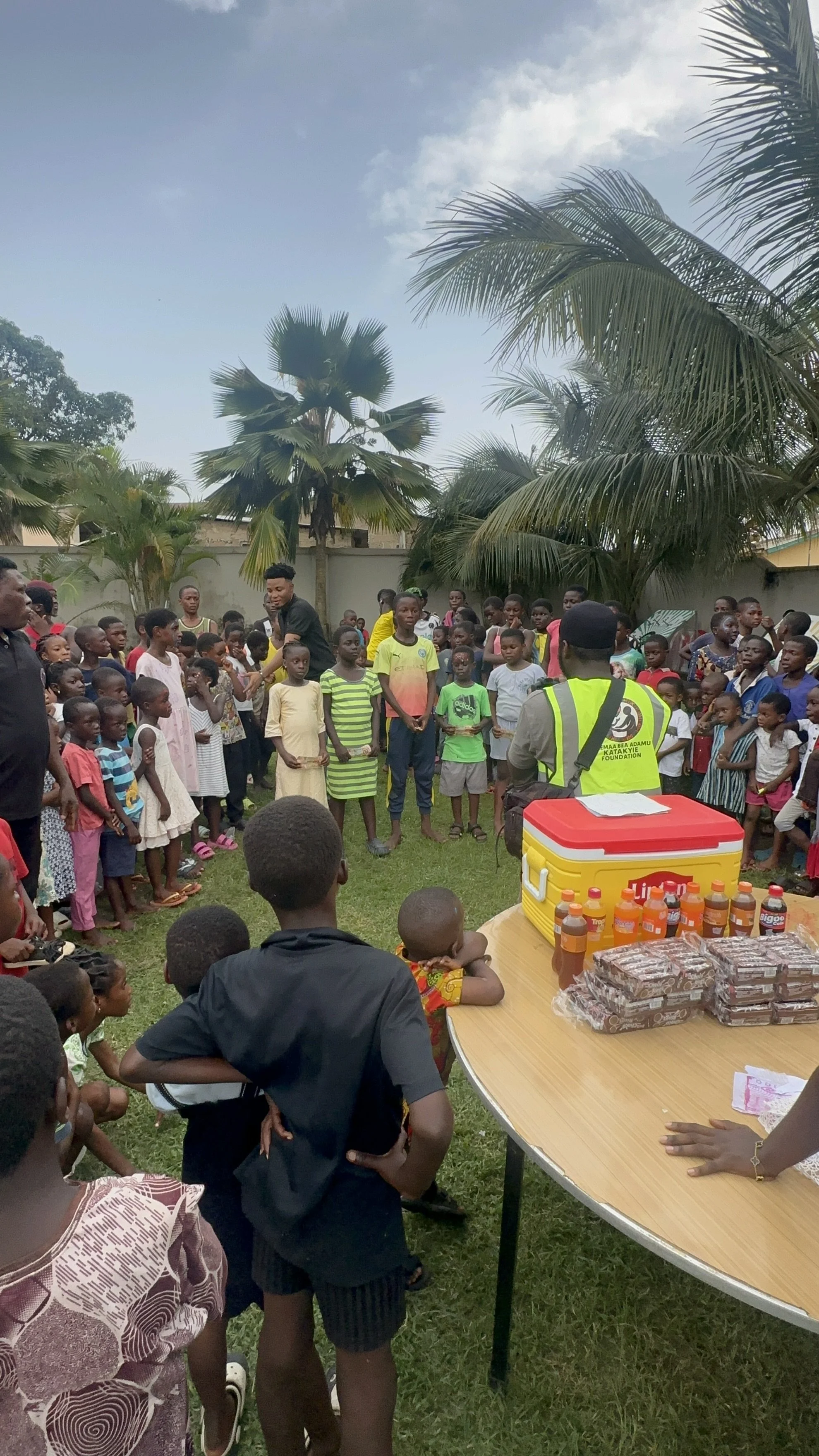 A group of children and a few adults gathered outdoors in a grassy area with palm trees, participating in a community event. There are tables with snacks and drinks visible in the foreground, including bottled beverages and wrapped packages, with one