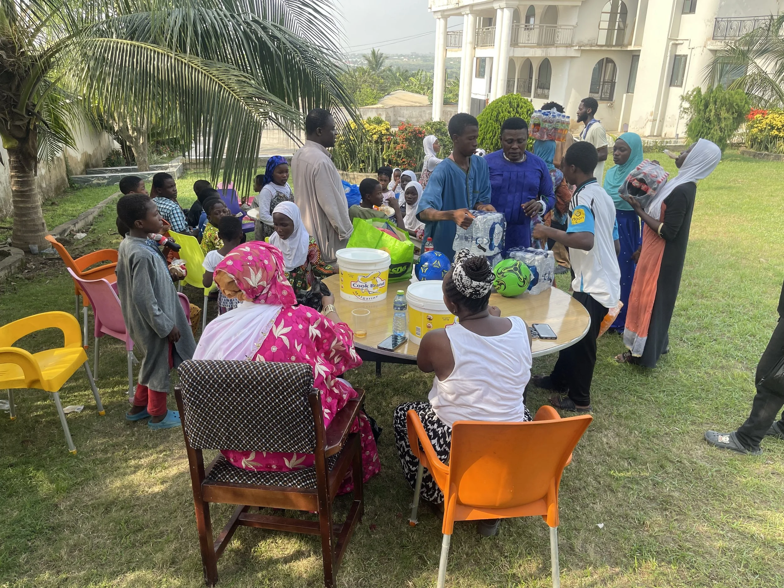 A group of people, including children and adults, gather outdoors around a table that has on it drinks and a bucket. People are engaging in conversations and some are handing out items. The setting appears to be a garden or backyard with a grass lawn