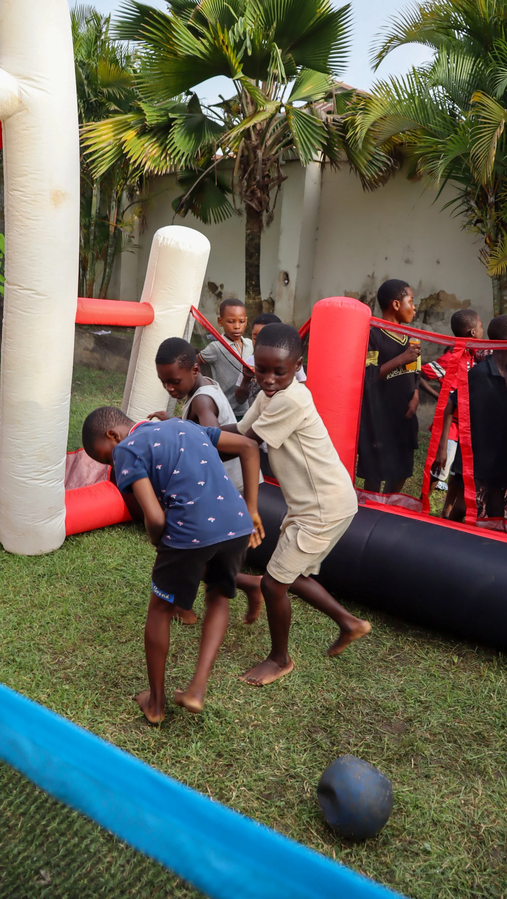 Children playing on a grass field with a red and black inflatable obstacle course, surrounded by a concrete wall and tropical trees.