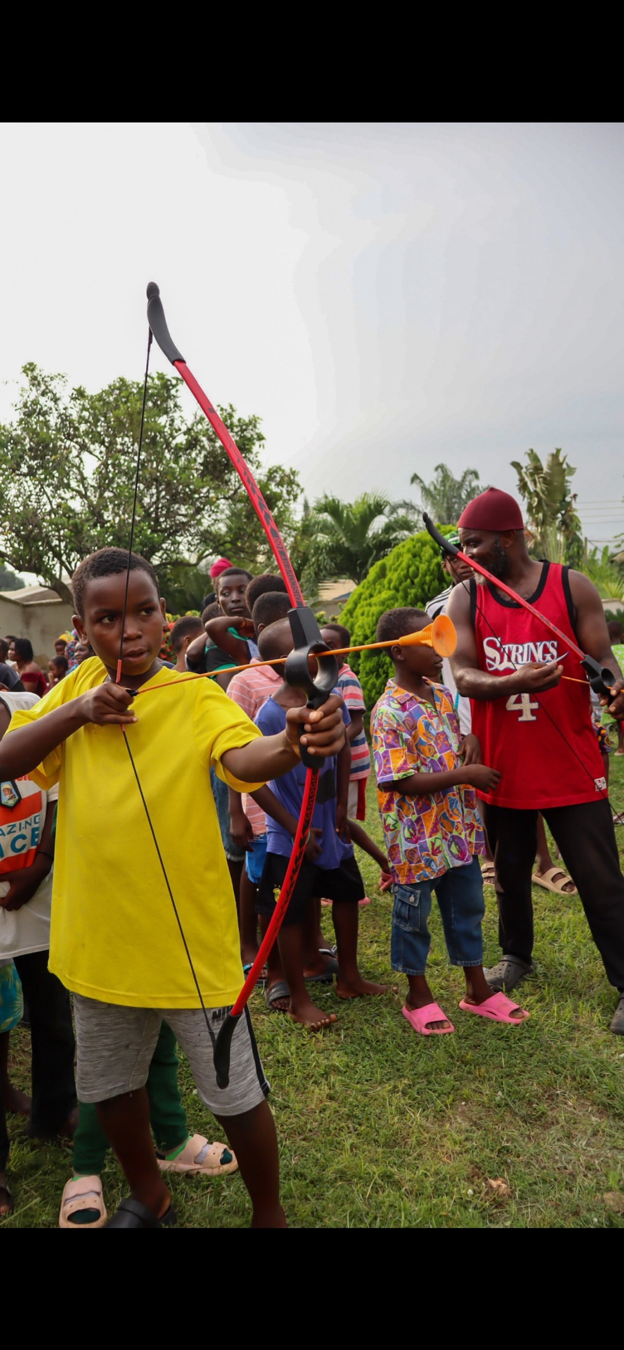 A young boy aiming a red and black archery bow with an orange arrow at a target during an outdoor event, surrounded by children and a man in a red tank top and red headscarf.
