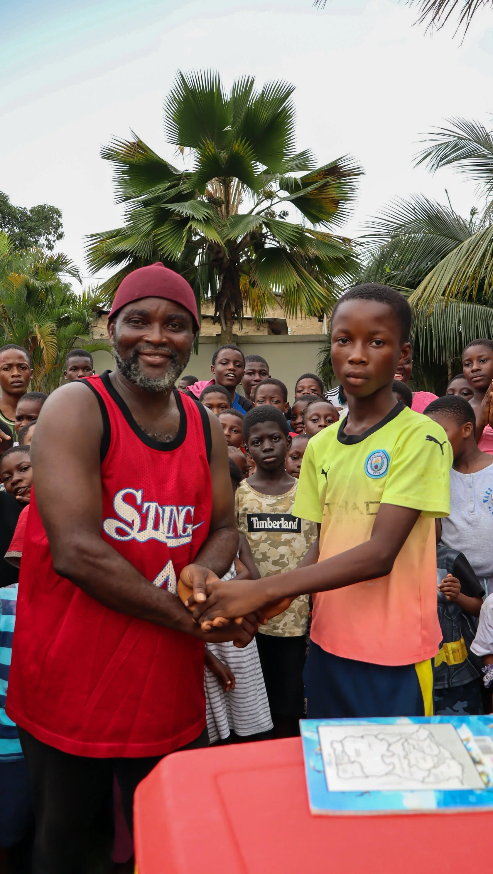 A man in a red sports jersey shaking hands with a young boy in a yellow and pink shirt among a group of children outdoors, with palm trees in the background.