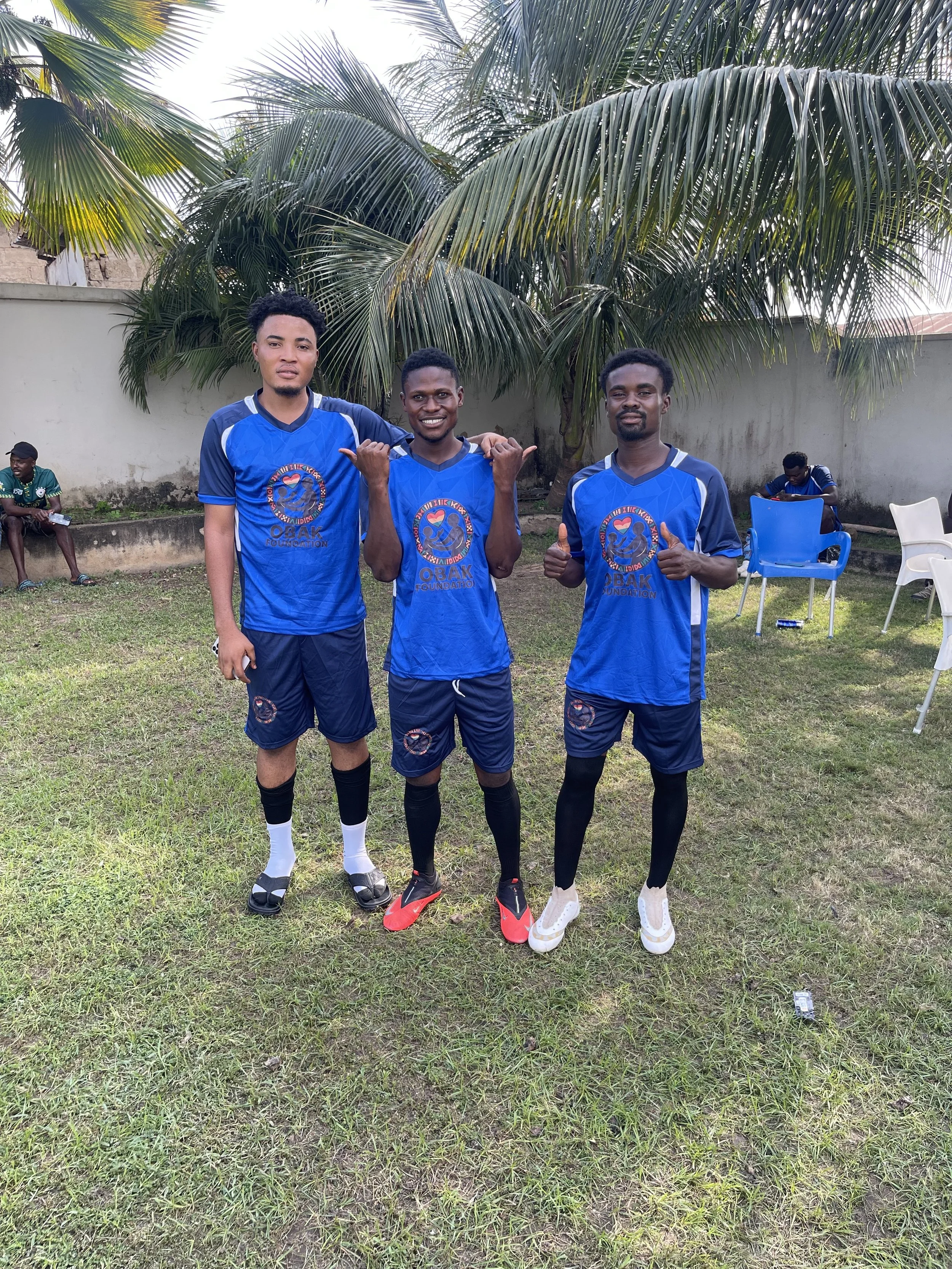 Three young men in blue sports jerseys and shorts standing on grass field, smiling and posing with thumbs up. Two of them are wearing black compression stockings, and one is wearing white shoes. Behind them, there are palm trees, a white wall, and a 