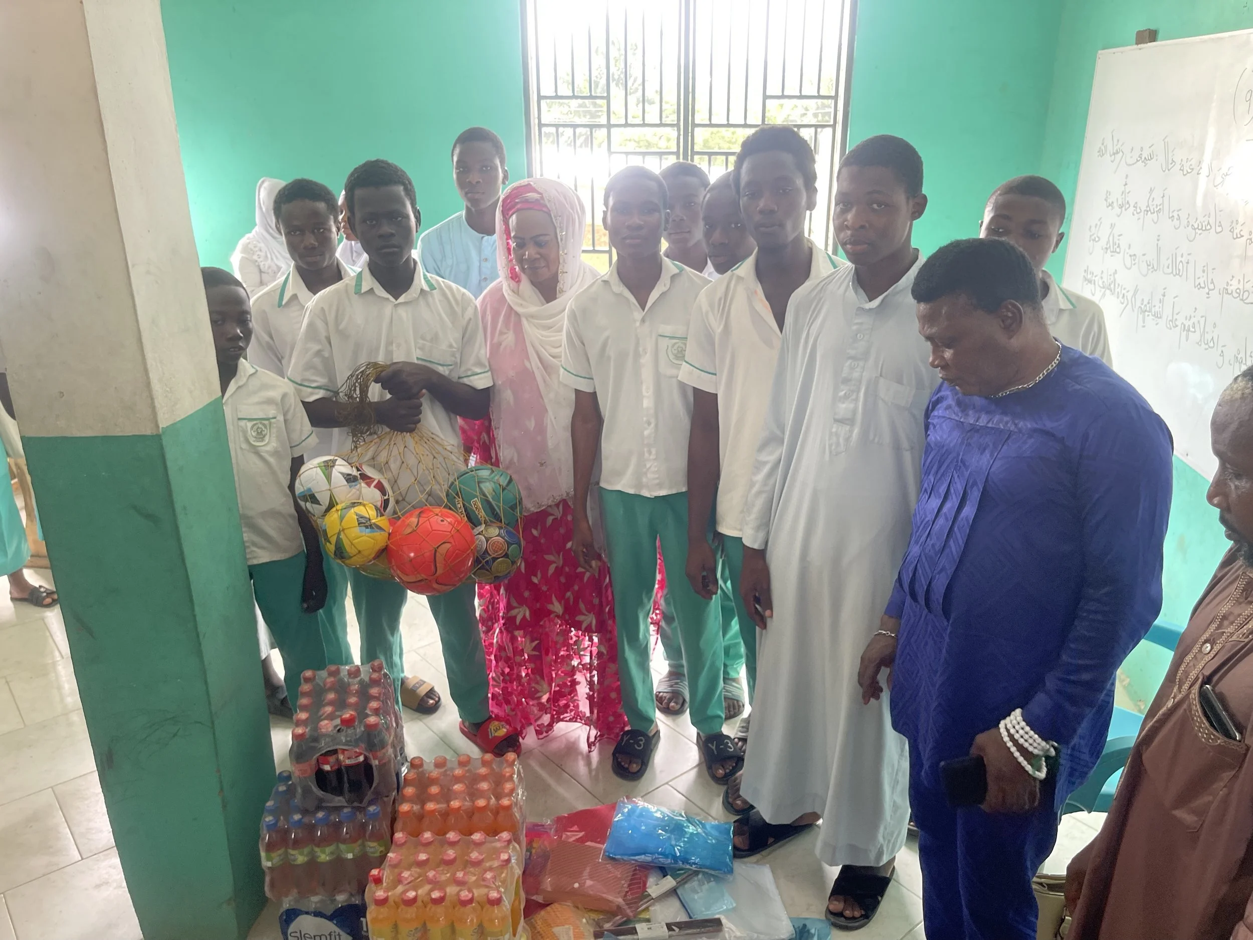 Group of students and adults gathered around supplies, including bottled drinks and sports balls, in a classroom with green walls and a whiteboard with writing.