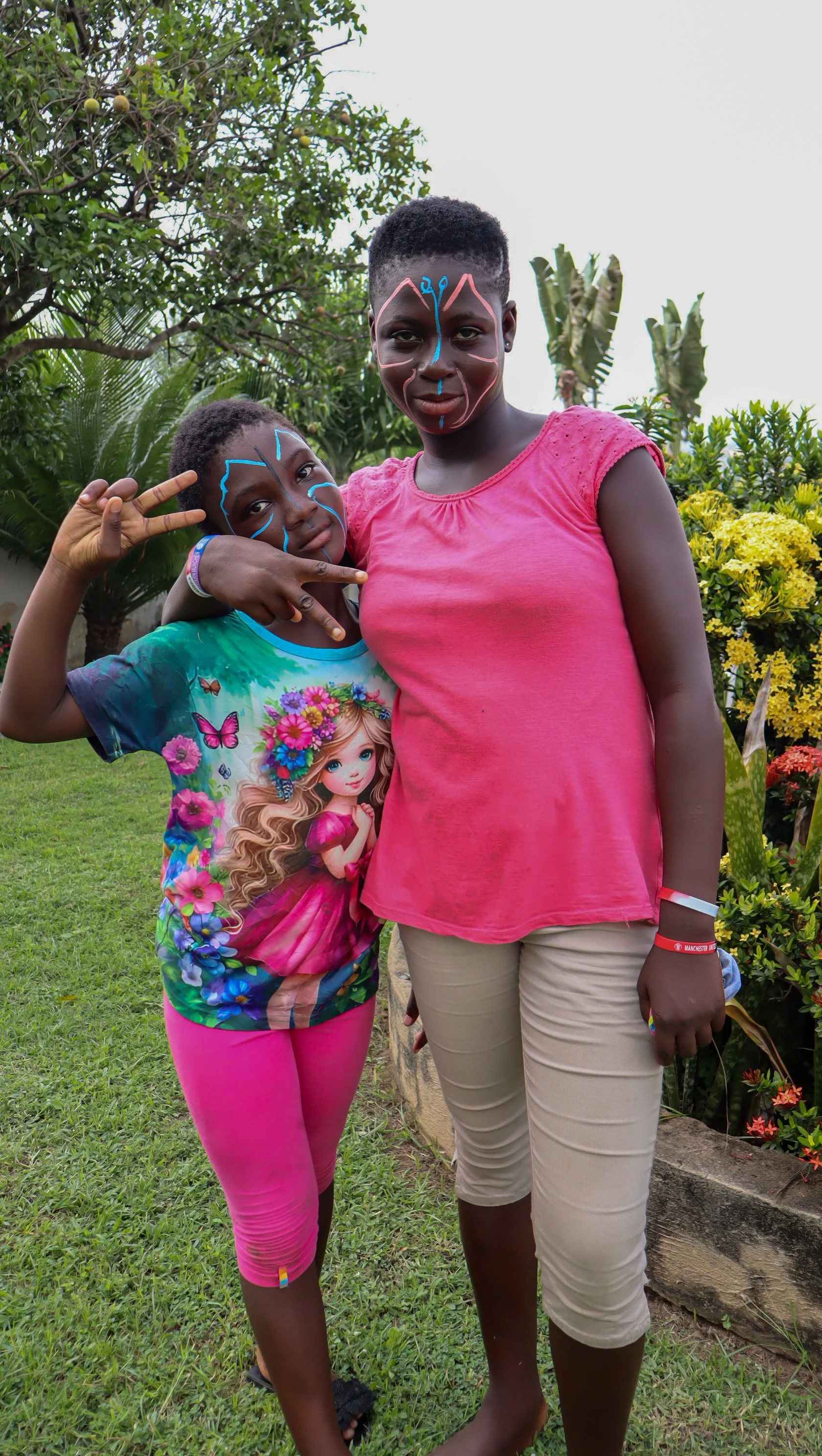 A young girl and an older girl with face paint, standing outdoors in a garden with trees and yellow flowers, smiling at the camera.