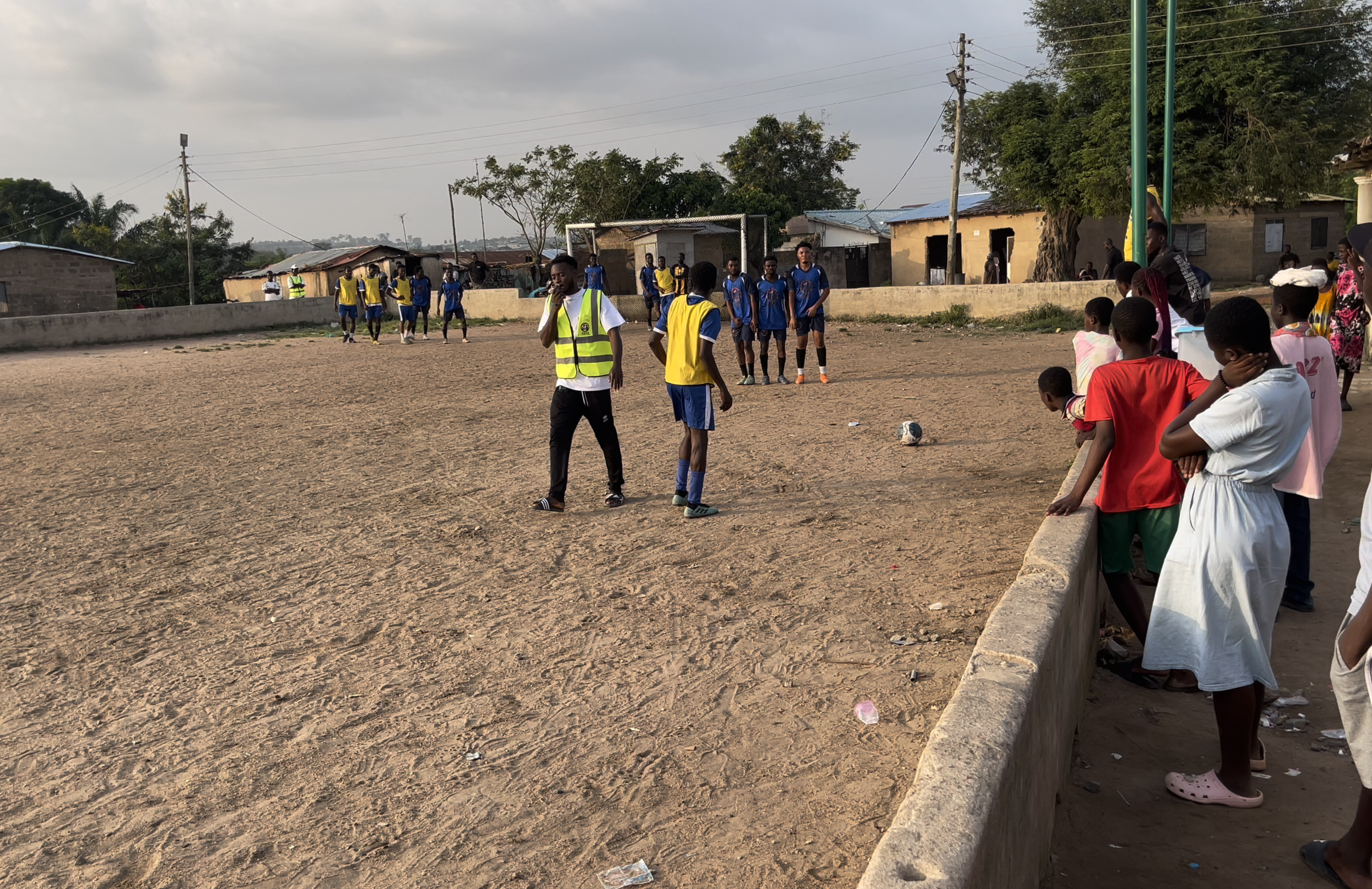 Children and teenagers watching a soccer game on a dirt field in a rural area with small buildings and trees in the background. Some players are dressed in blue jerseys, and one person in a reflective vest is standing near the field.