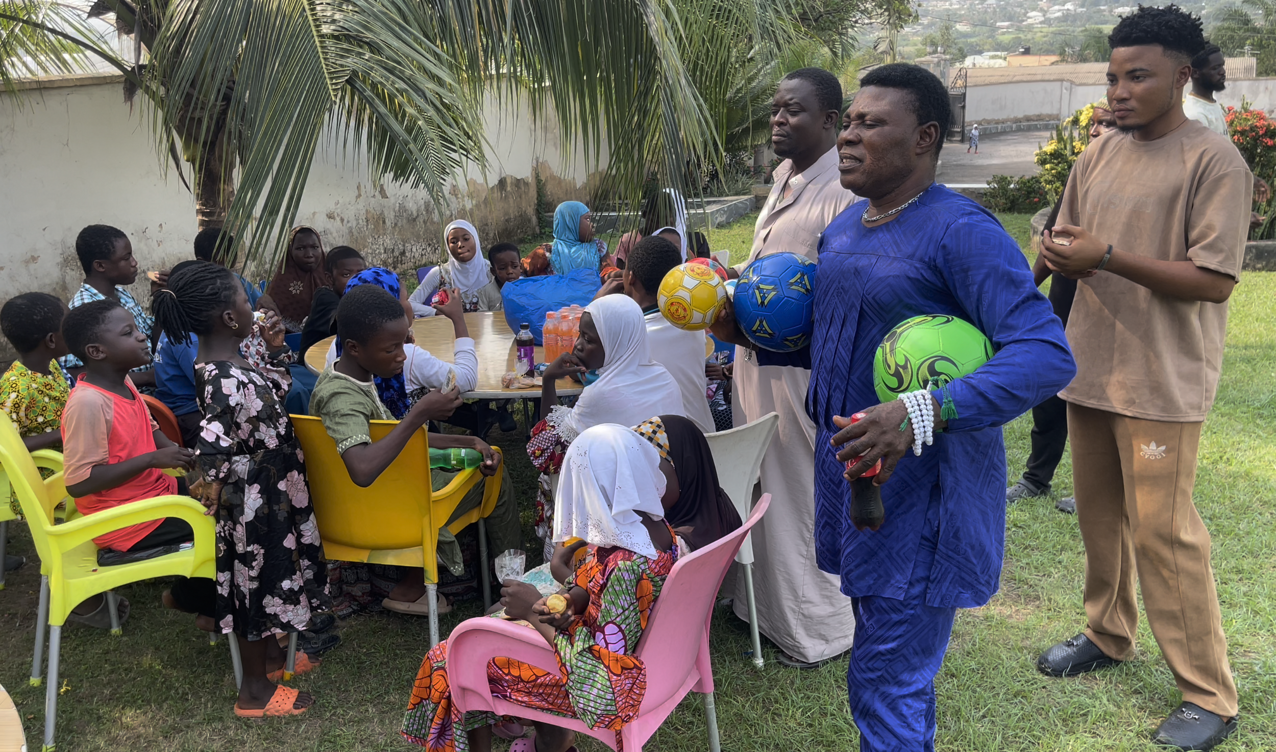 People gathered outdoors around a table, with some holding soccer balls and various drinks, children seated and standing, engaging in what appears to be a celebration or gathering in a grassy area with trees and a wall in the background.