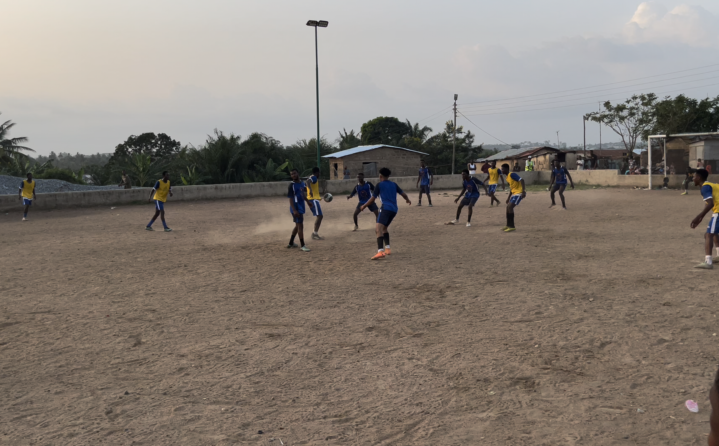 A group of young men playing soccer on a dirt field with some houses and trees in the background during daytime.