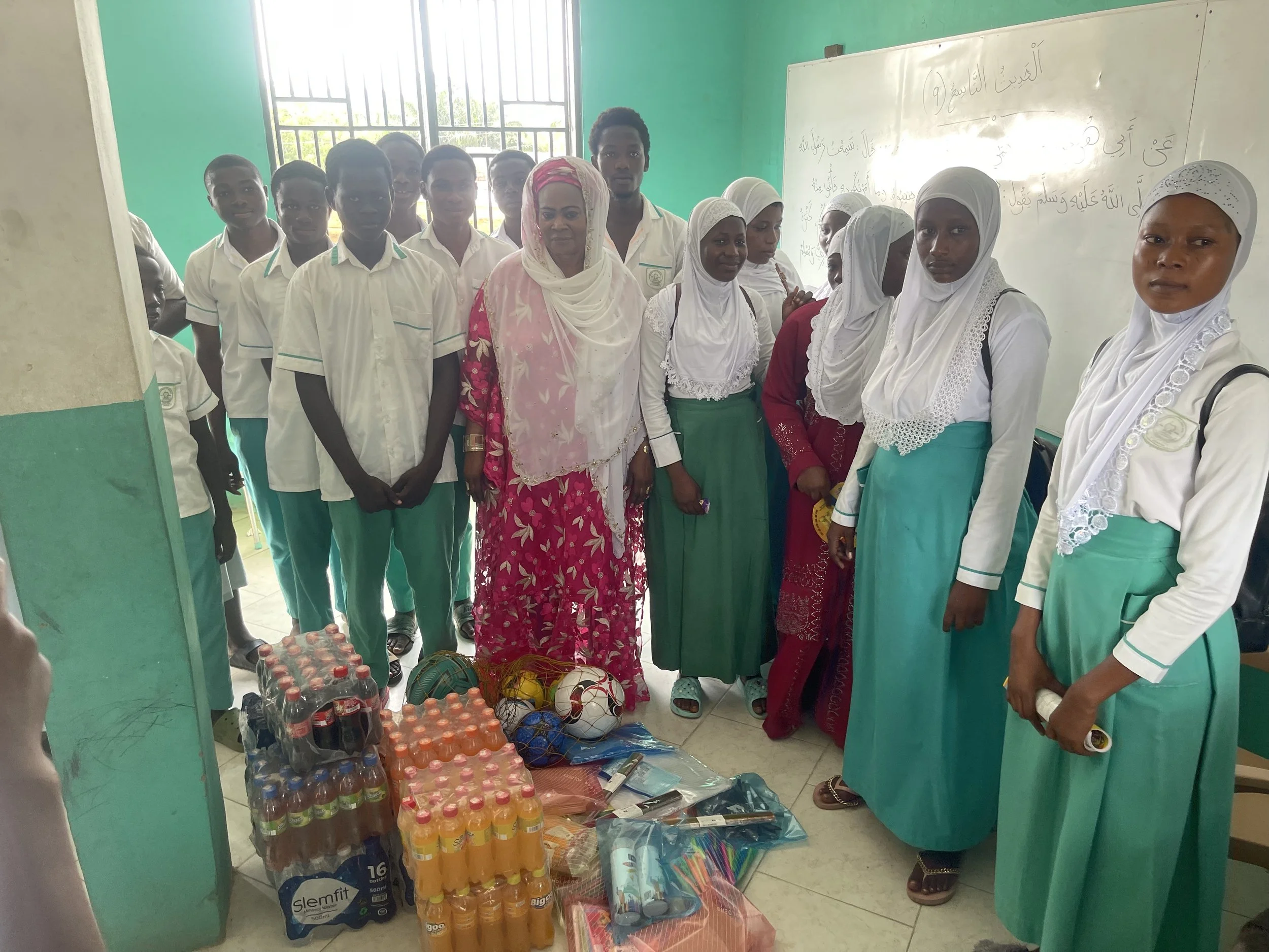 Group of students and teachers in a classroom with supplies including bottled drinks, sports balls, and school materials on the floor.
