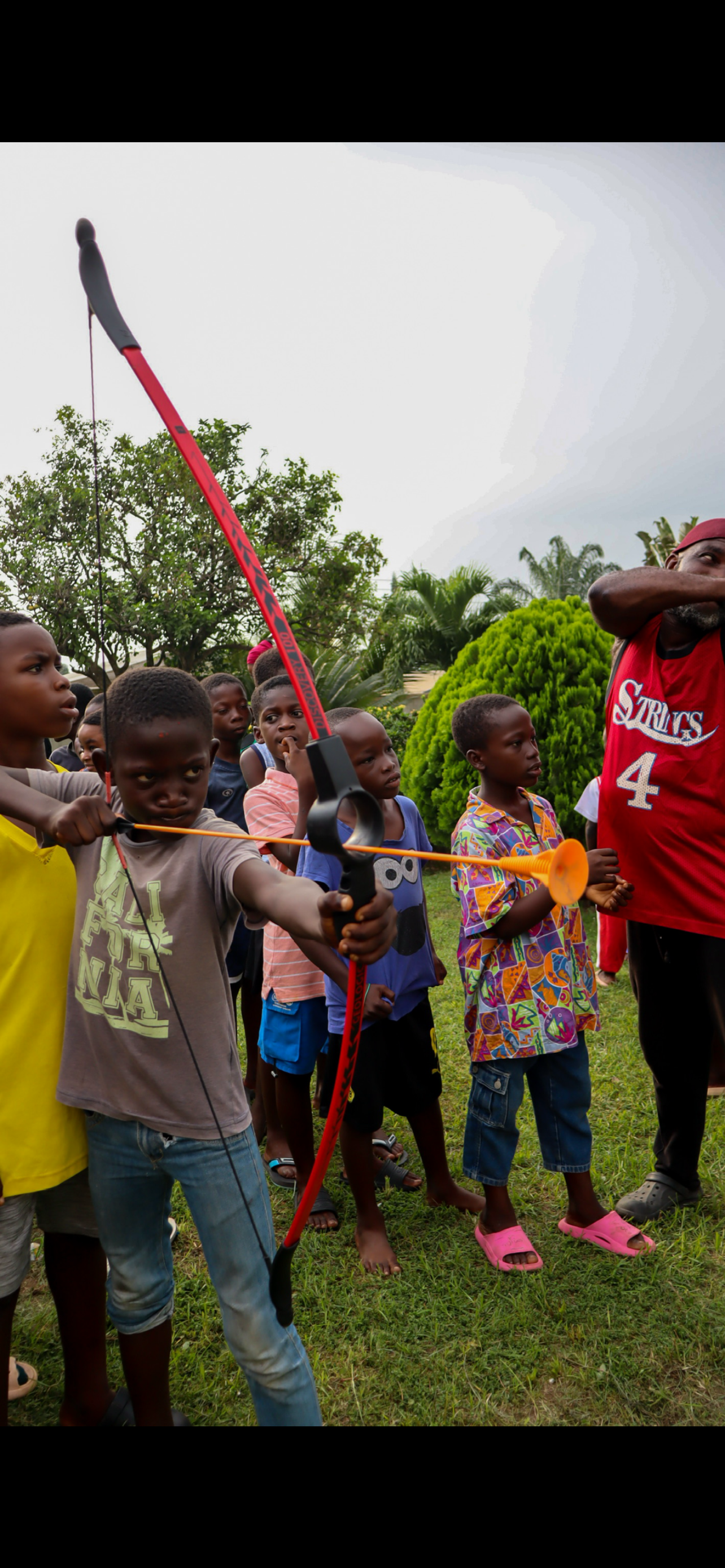 Children participating in an outdoor archery activity, with one child drawing a bow and aiming an arrow, surrounded by other children watching attentively.