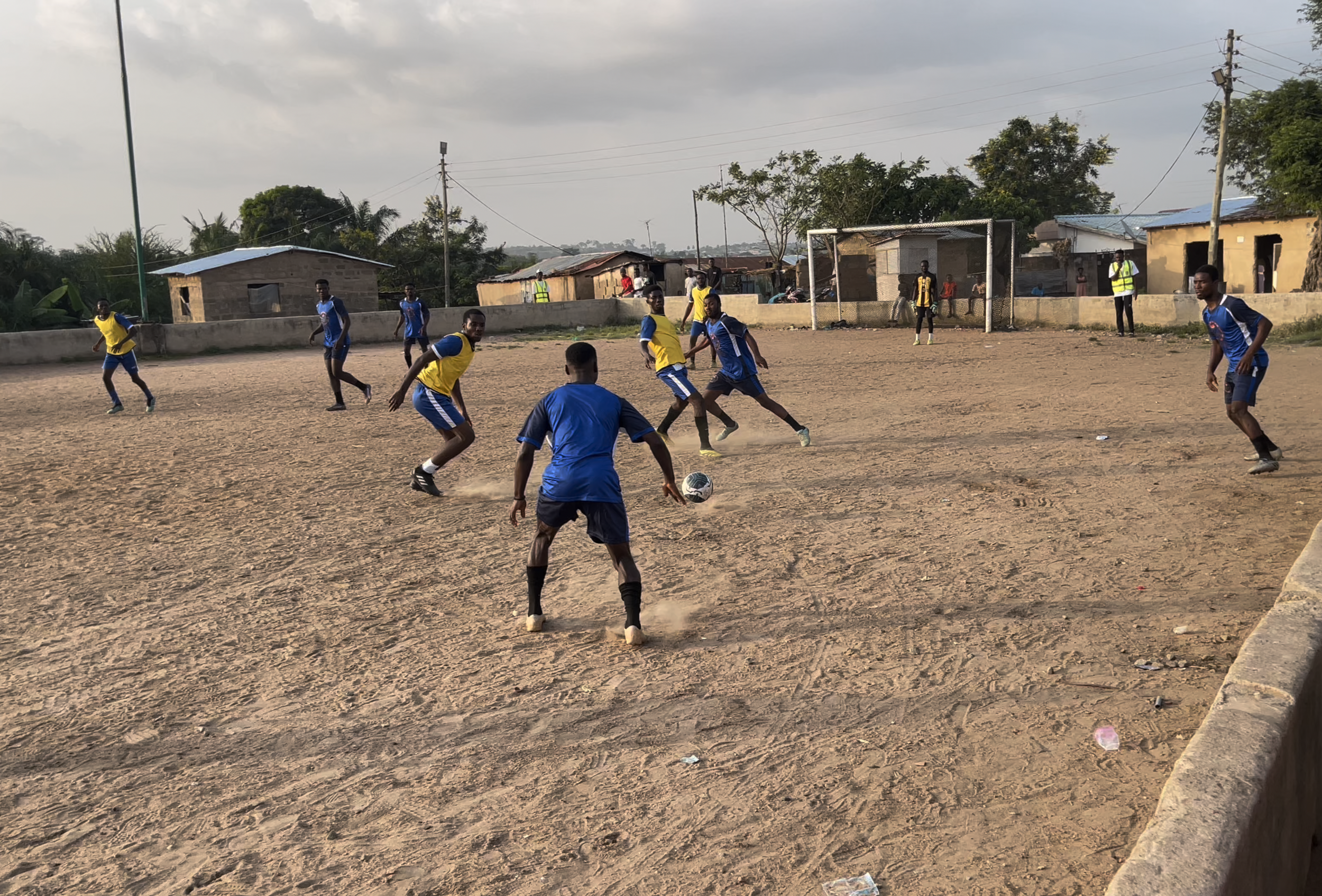 A group of young men playing soccer on a dirt field, some wearing blue jerseys, others in yellow, with houses and utility poles in the background.