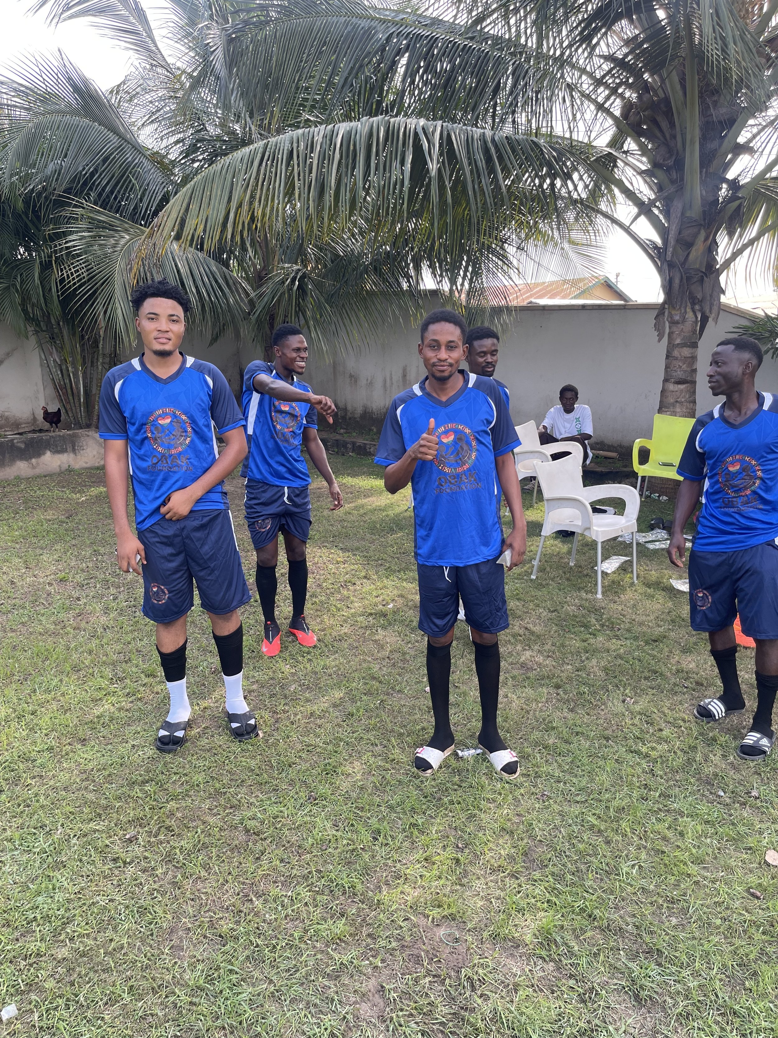 A group of five young men in matching blue soccer uniforms standing on a grassy field with palm trees, white chairs, and a concrete wall in the background. One man is giving a thumbs up.