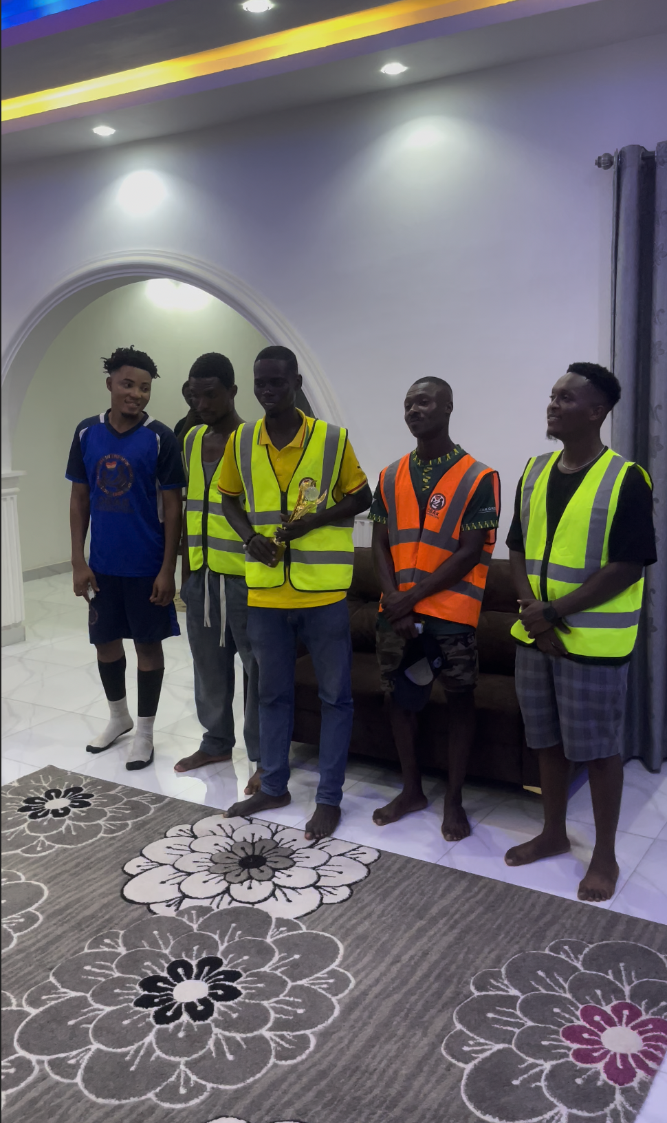 Five young men standing indoors on a tiled floor in front of a white wall with a curved archway and a floral carpet. Four of the men are wearing high-visibility safety vests, one of them is holding a trophy.