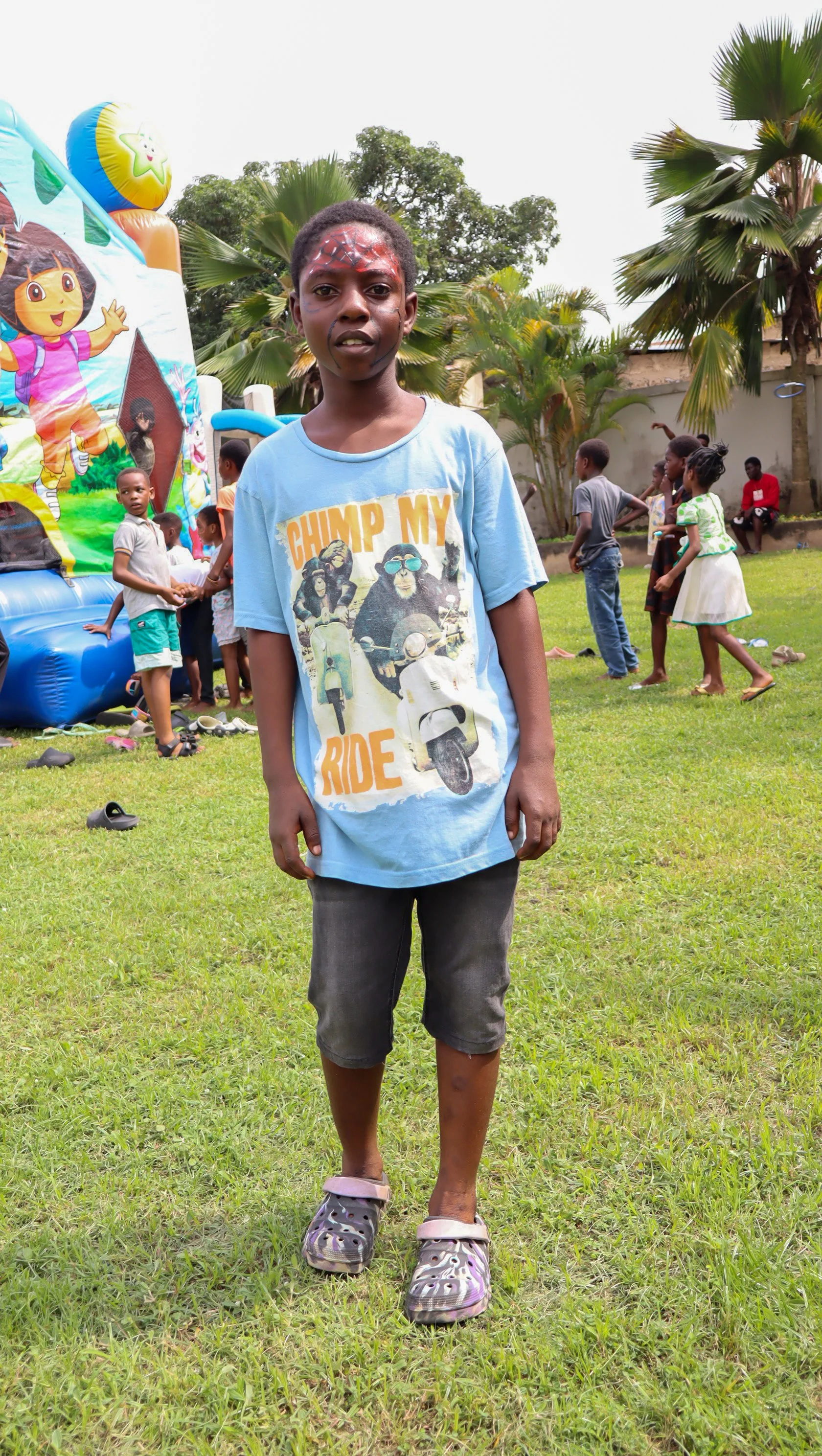 A young boy standing on a grassy field, wearing a blue t-shirt with a graphic of monkeys and motorcycles, gray shorts, and purple Crocs. In the background, children are playing near an inflatable bounce house with colorful cartoon characters, surroun