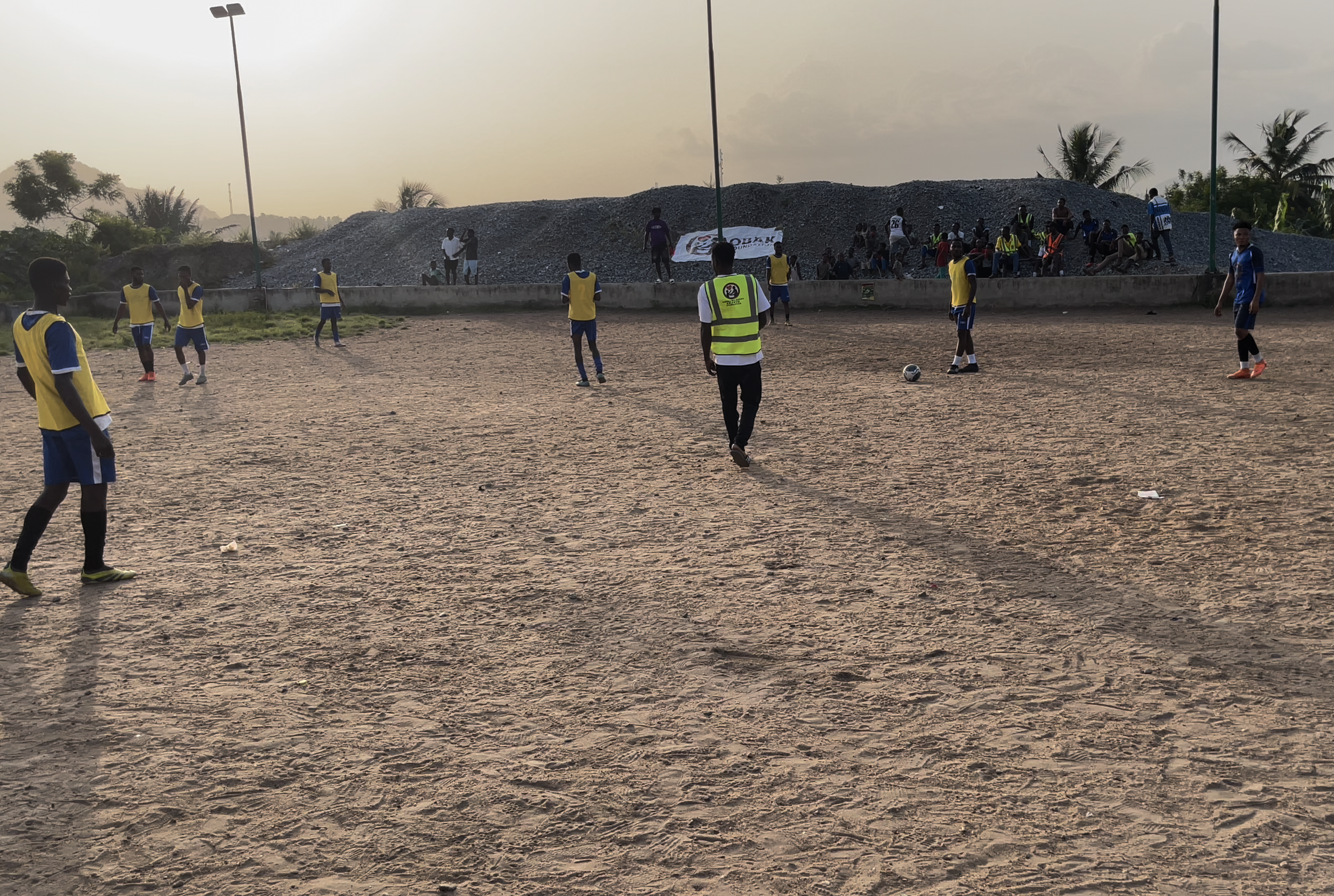 A group of young men playing soccer on a dirt field with some spectators sitting on the sidelines in the background during sunset.