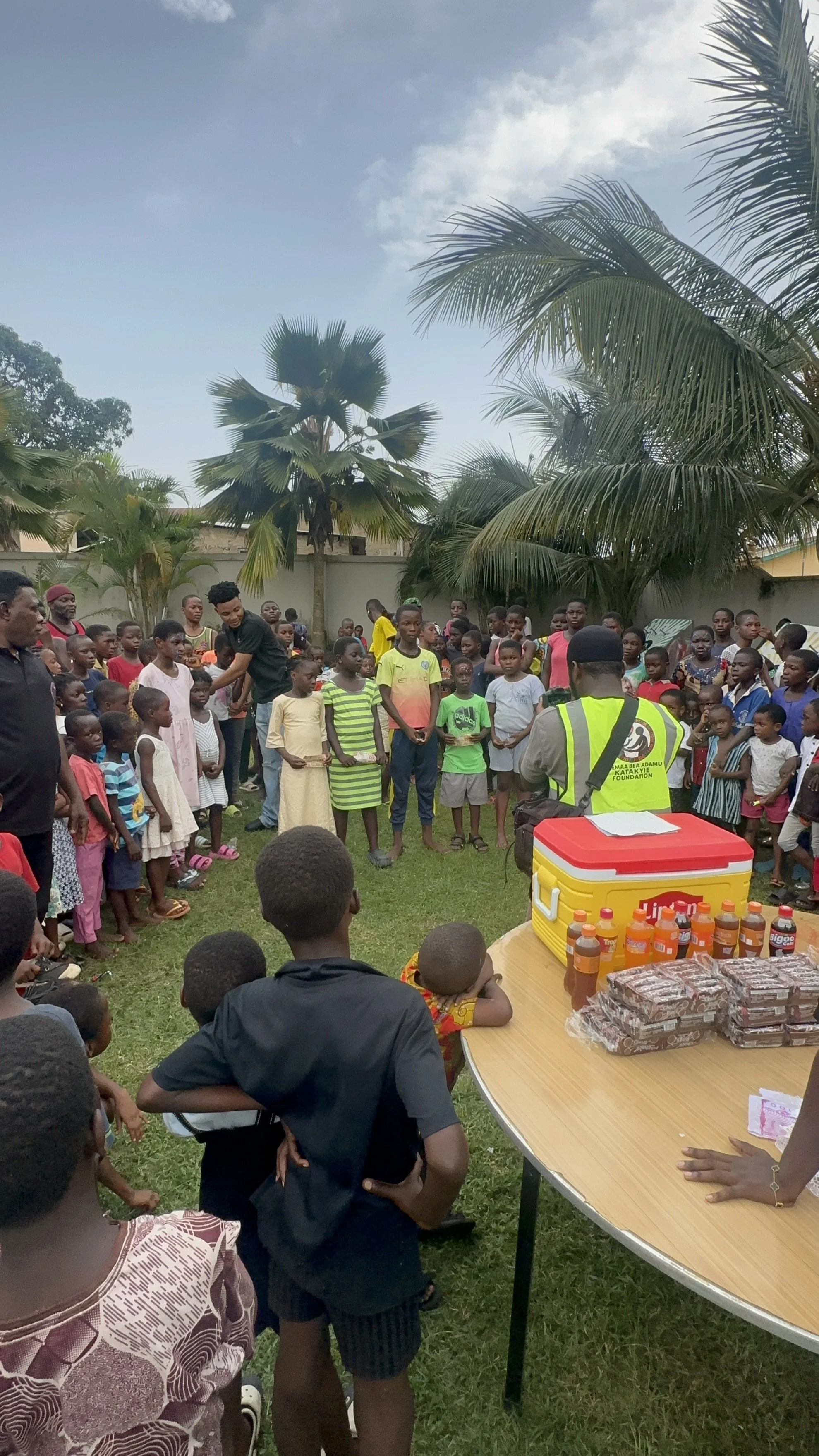 Children and adults gathered outdoors around a table with drinks and snacks, with trees and a fence in the background, during a community event or celebration.