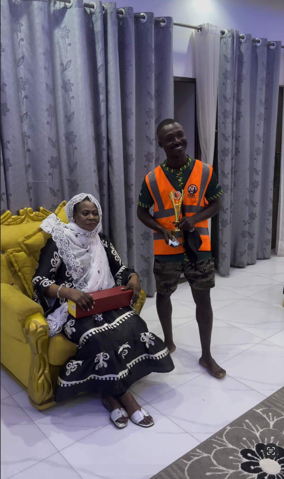 A man in a bright orange safety vest holding a trophy, standing next to a seated woman dressed in black and white traditional attire with a hijab, holding a red box, in a room with gray curtains and a yellow armchair.