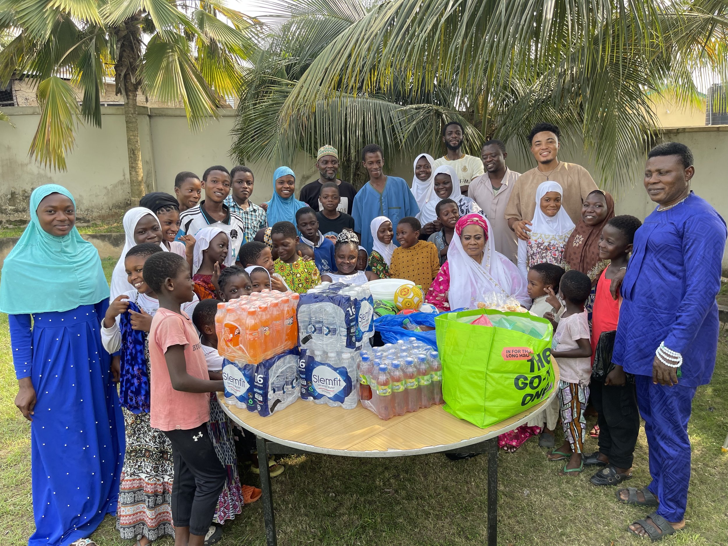 A large group of people, including children and adults, gathered outdoors around a table with drinks and gifts. Palm trees and a wall are in the background, indicating a tropical setting. The group appears to be celebrating a special occasion.