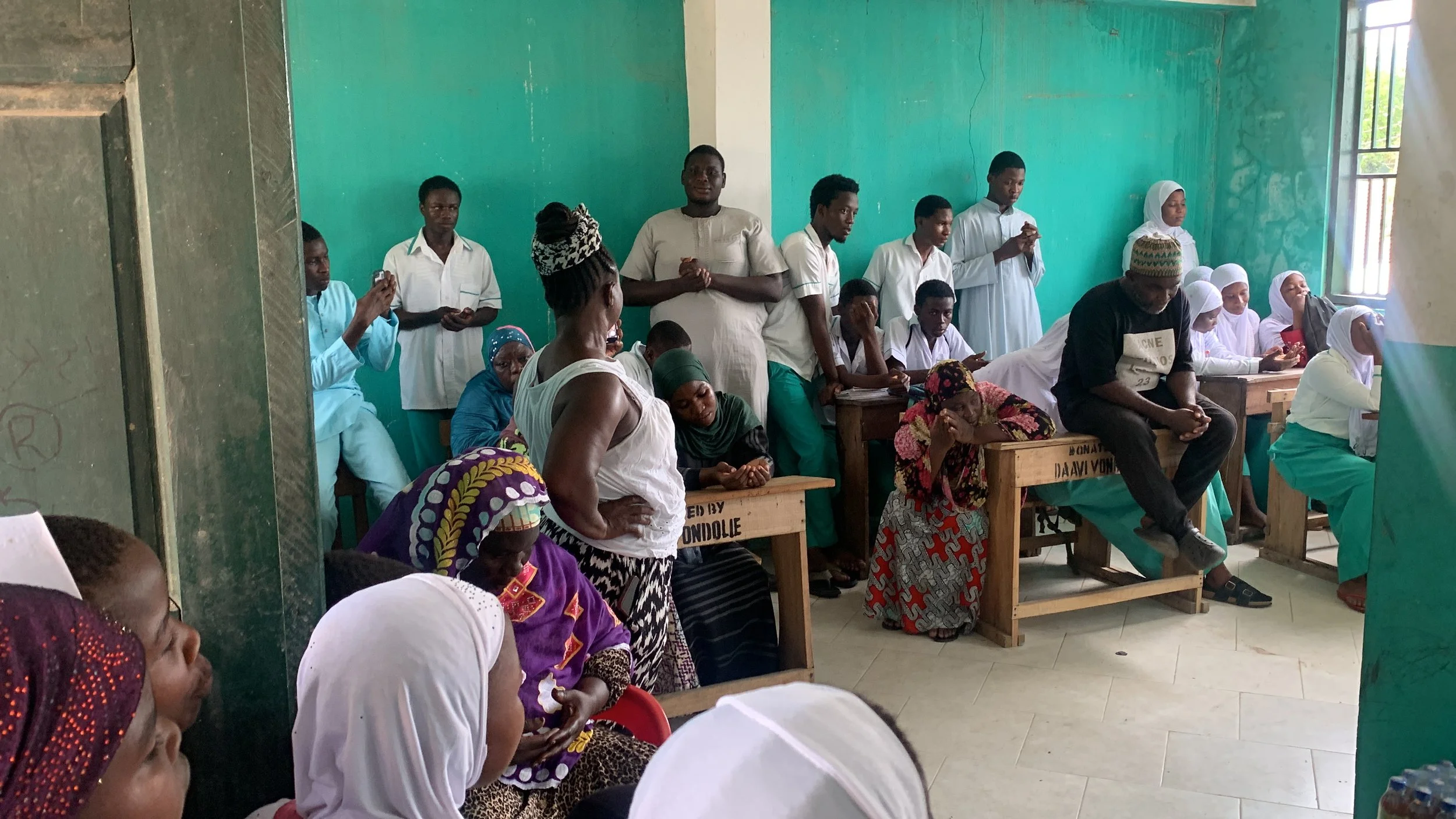 Group of people gathered inside a room with teal walls, some seated and some standing, engaged in prayer or reflection, with sunlight coming through the window on the right.