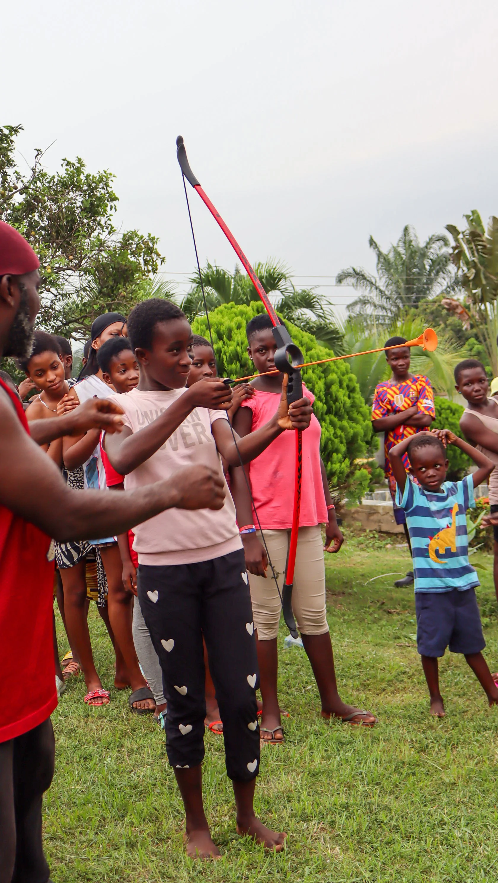 A boy practicing archery outdoors with several children and an adult watching, in a grassy area with trees and a cloudy sky.