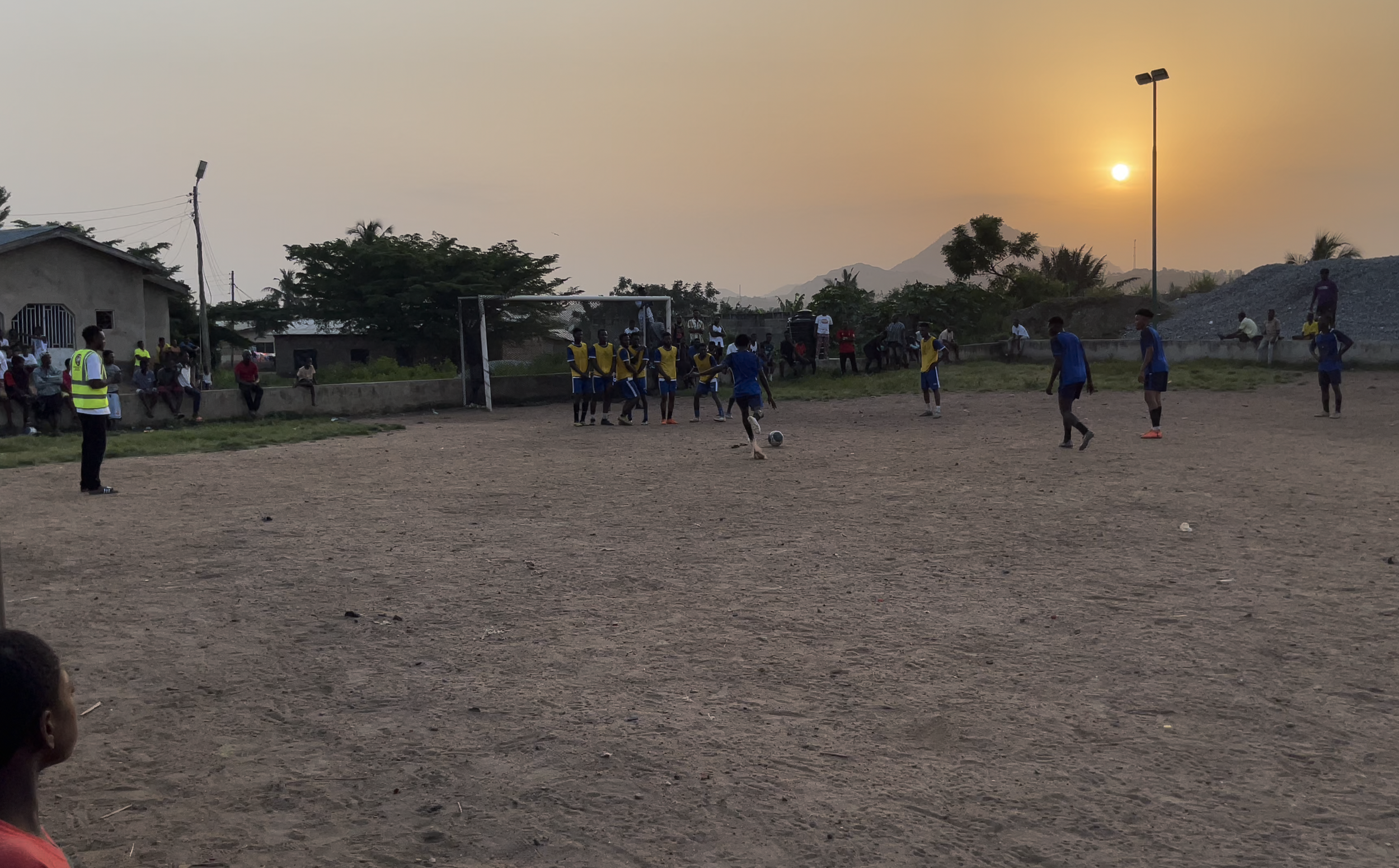 Young soccer players practicing on a dirt field during sunset, with spectators sitting on a concrete wall and trees in the background.
