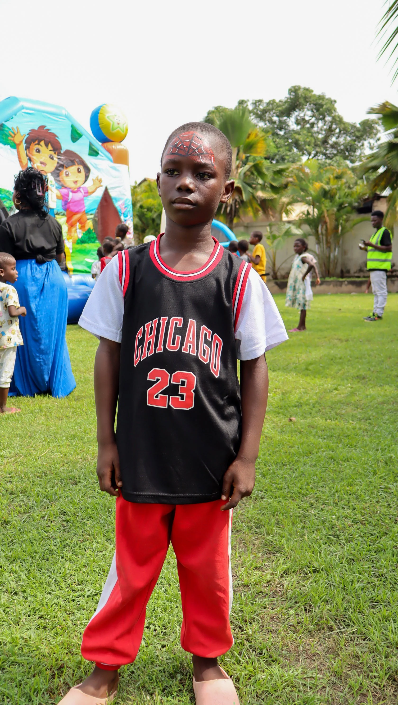 A young boy with face paint resembling a spider web wears a Chicago basketball jersey and red pants, standing on grass at an outdoor party with children, some balloons, and palm trees in the background.