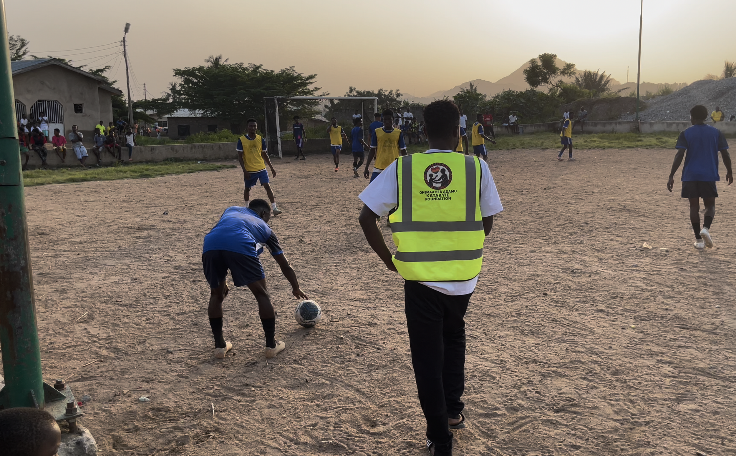 People playing soccer on a dirt field during sunset with spectators sitting on the sidelines and mountains in the background.
