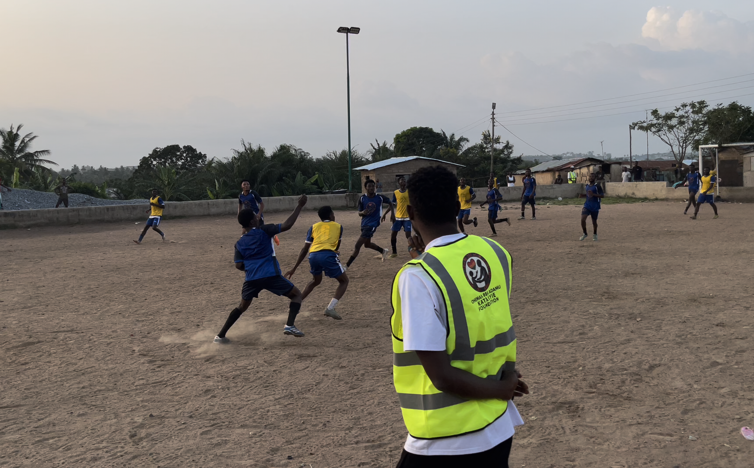 Children playing soccer on a dirt field, with spectators and small buildings in the background, and a person in a reflective vest observing.
