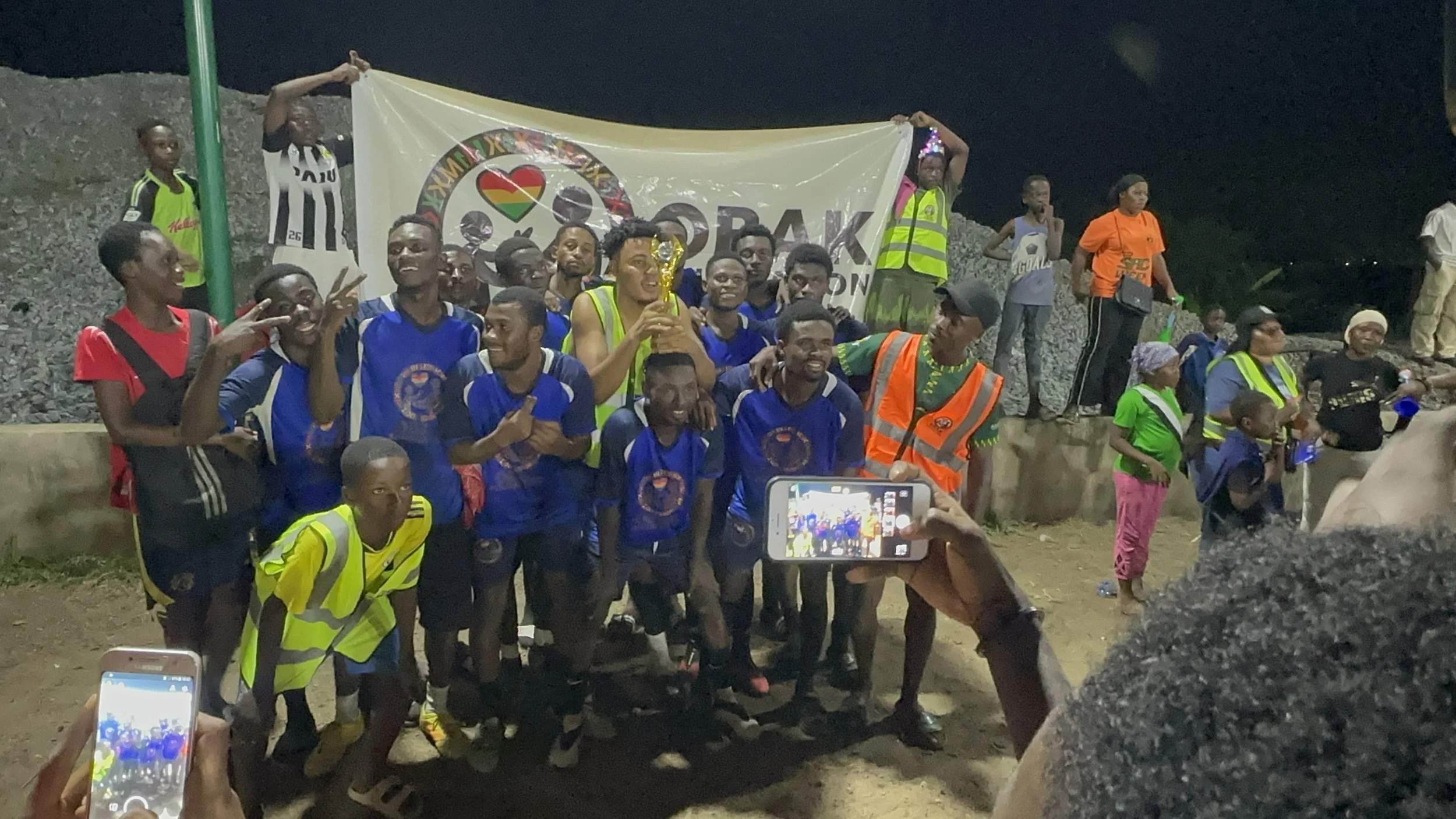 Group of people celebrating a sports victory at night, some holding a trophy, with a banner in the background, and others taking photos.