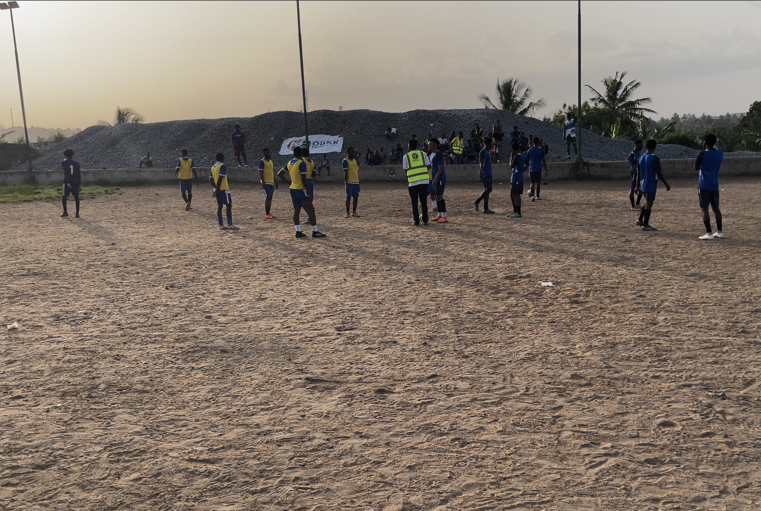 Soccer players in yellow and blue uniforms on a dirt field practicing with a coach or referee in a reflective vest, with spectators and palm trees in the background during sunset.