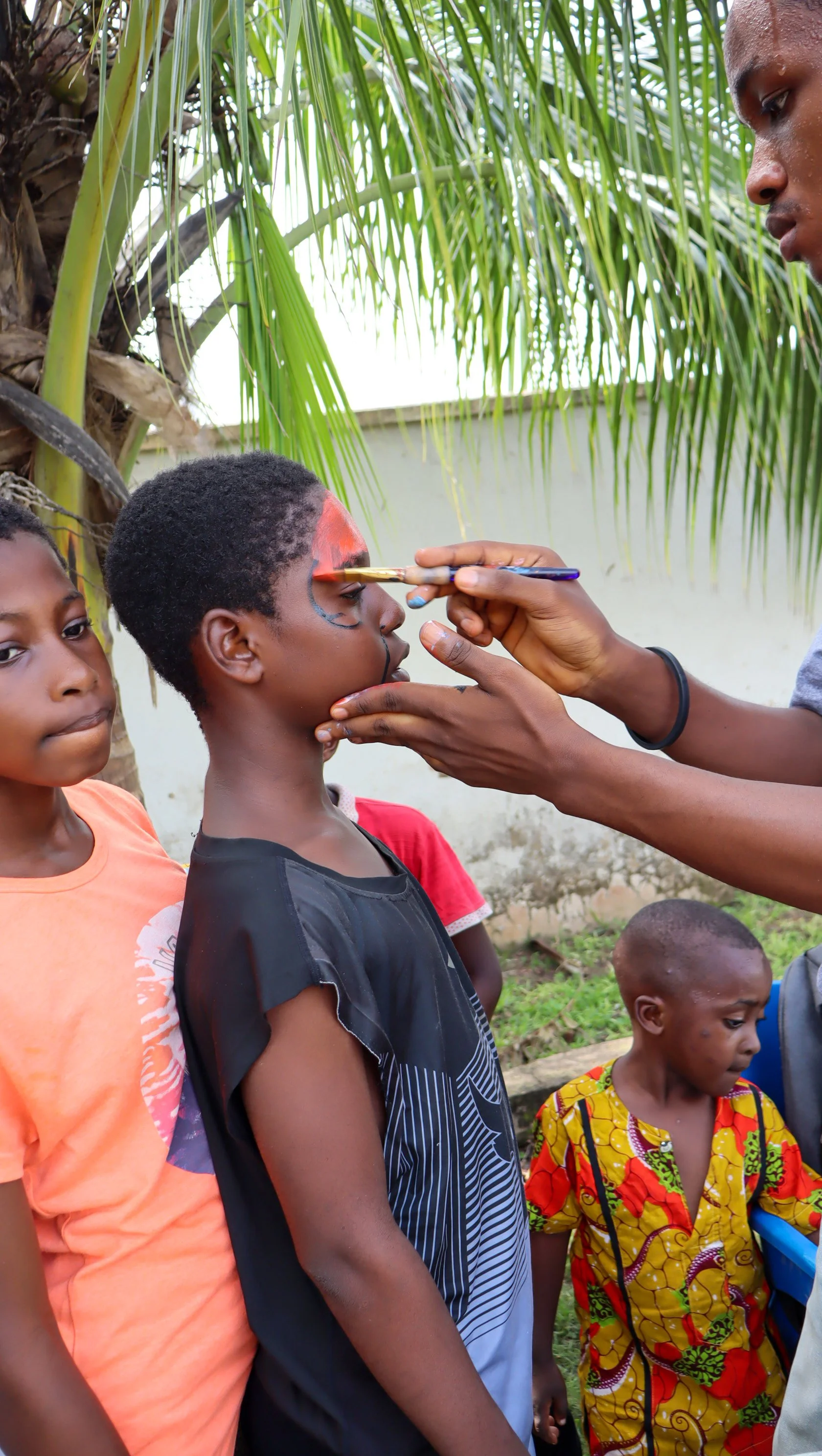 A young boy is getting face paint from an artist, surrounded by other children near a palm tree and a wall.