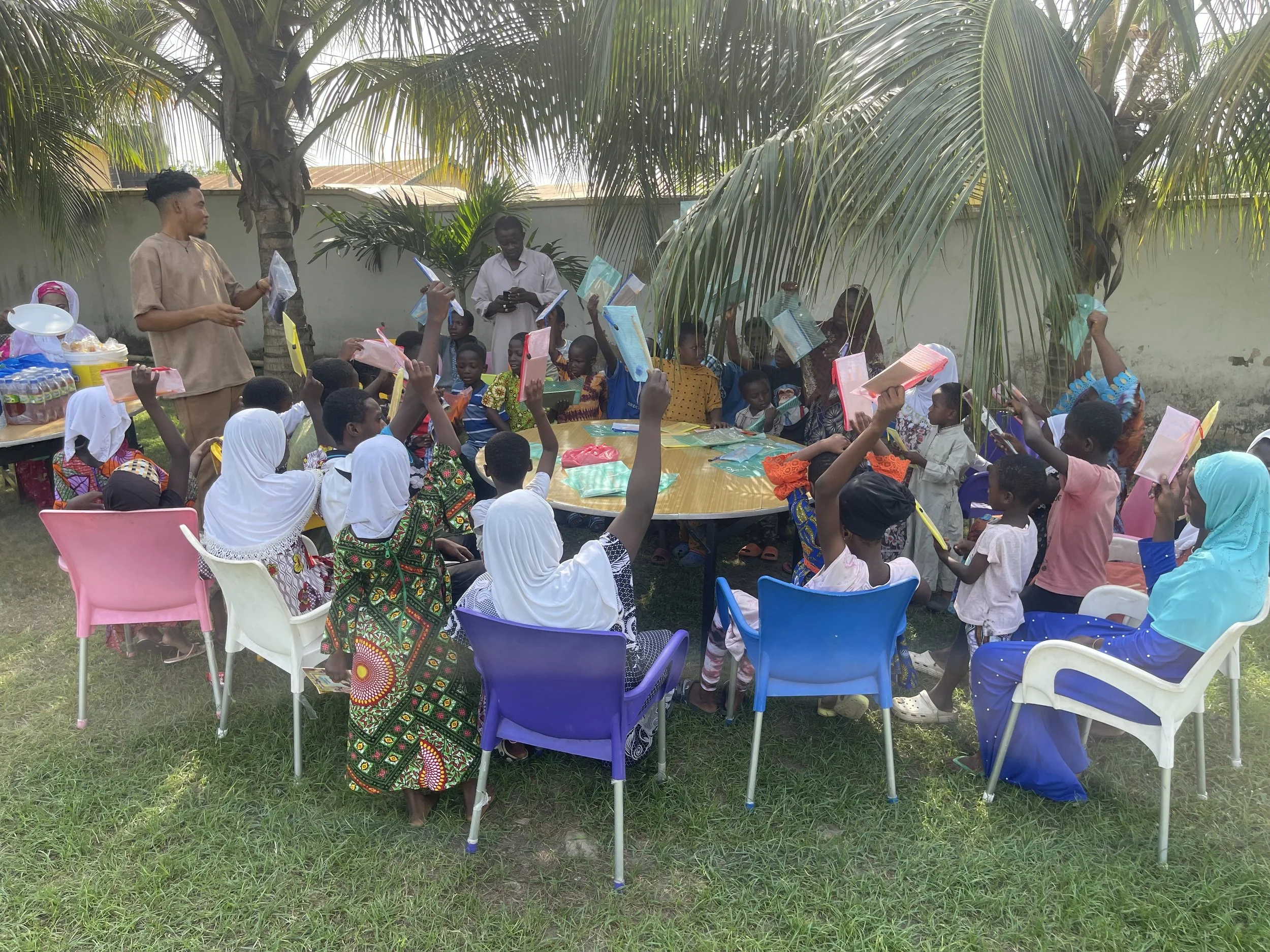 Group of children and adults gathered outdoors around a table, raising notebooks or folders in the air, with a palm tree and a wall in the background.