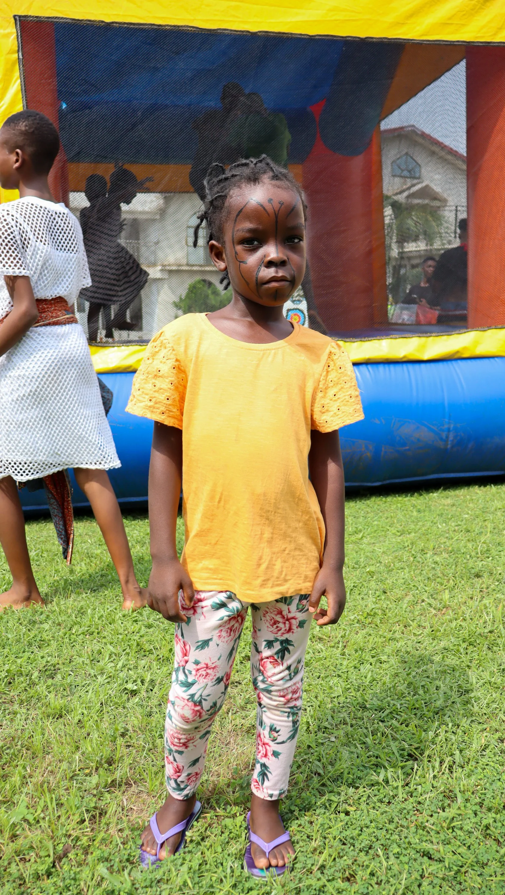 A young girl standing on grass with butterfly face paint, wearing a yellow top, floral pants, and purple flip-flops, in front of a bounce house at an outdoor event.