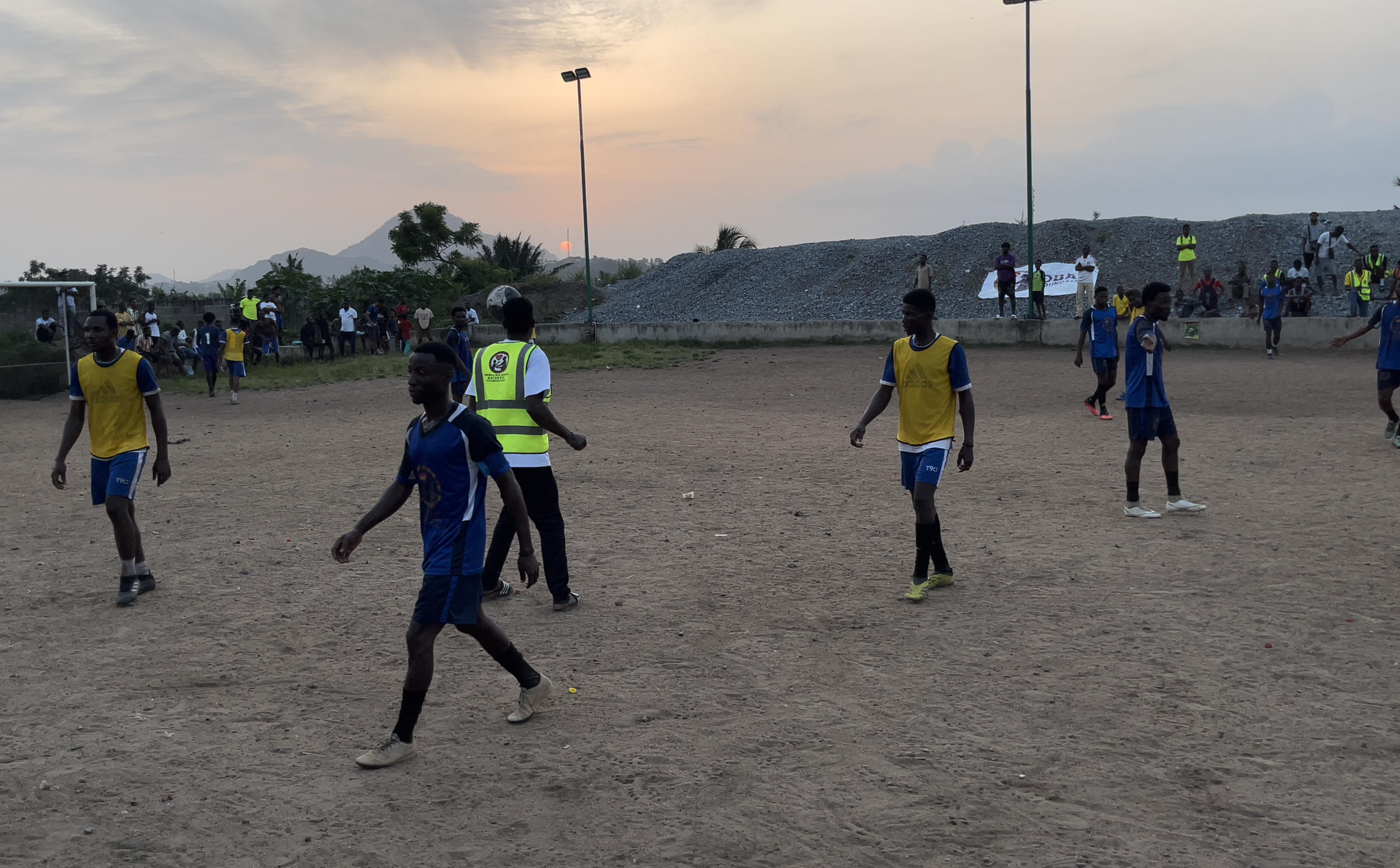 Young soccer players in blue and yellow jerseys playing on a dirt field during sunset, with spectators and officials in the background.