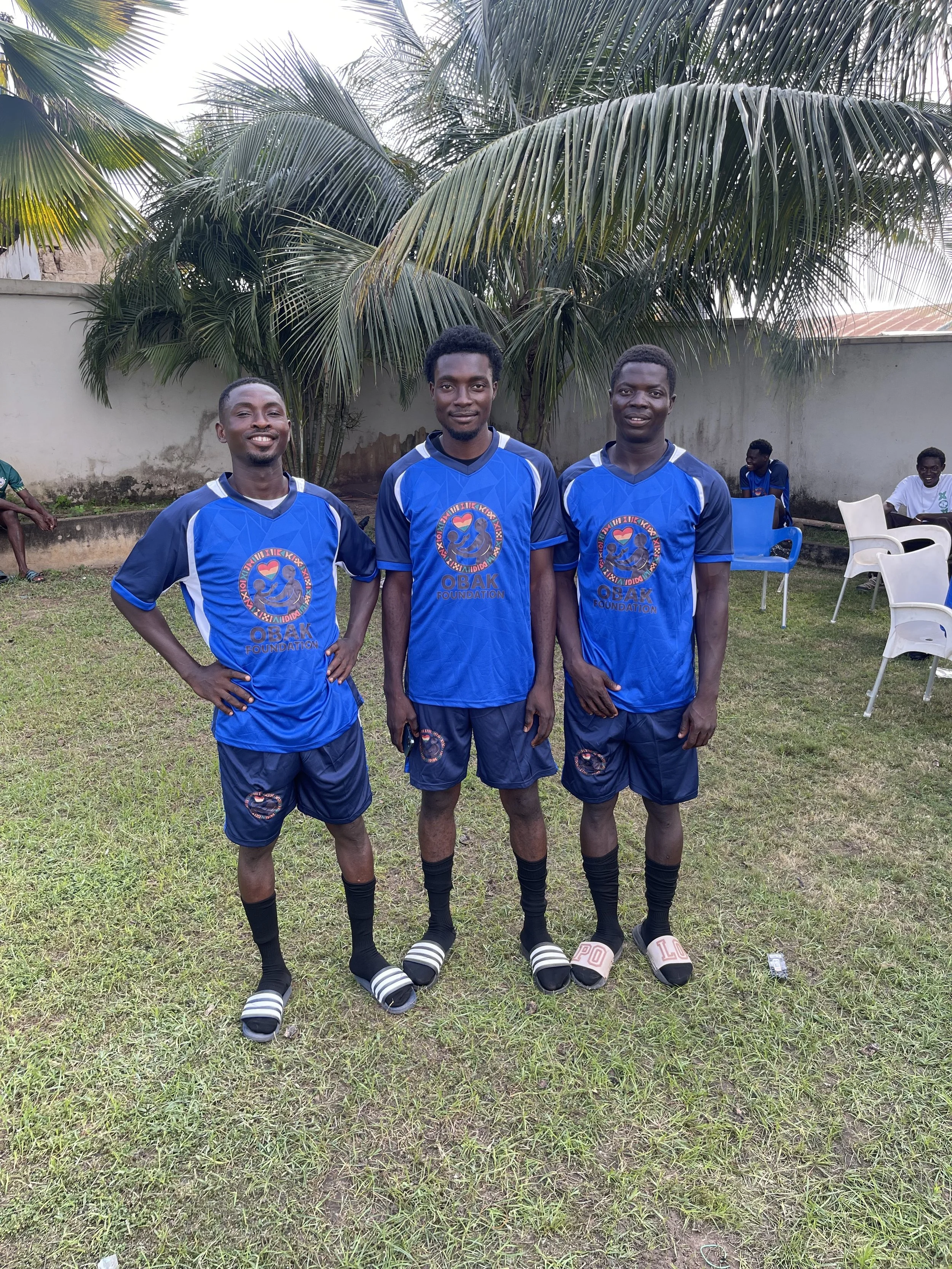 Three young men in blue soccer jerseys and shorts standing on grass with palm trees and chairs in the background.