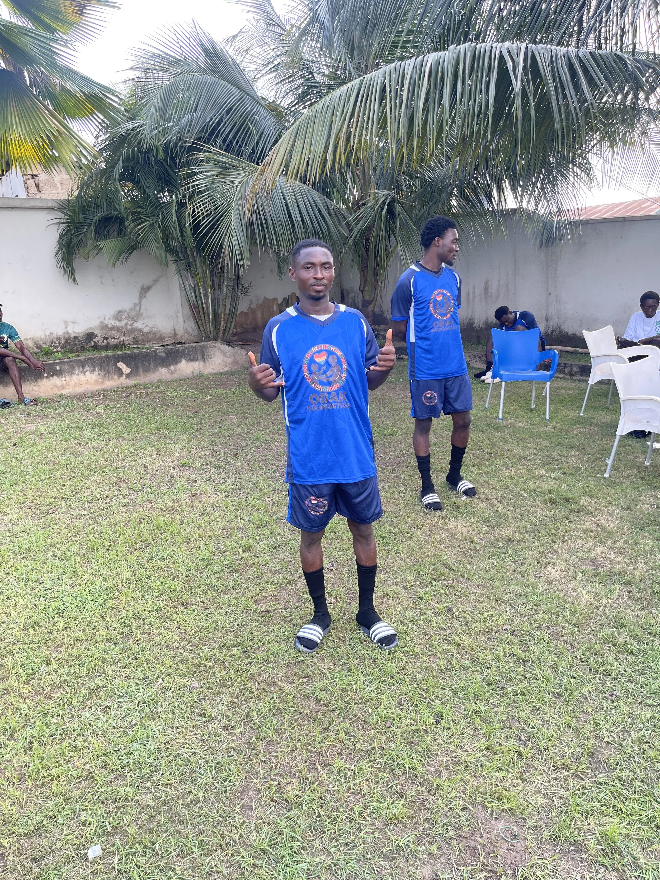 Two young men in blue soccer uniforms standing on a grassy field with palm trees and a concrete wall in the background; one is giving a thumbs up, the other is looking to the side. Several chairs and other people are in the background.