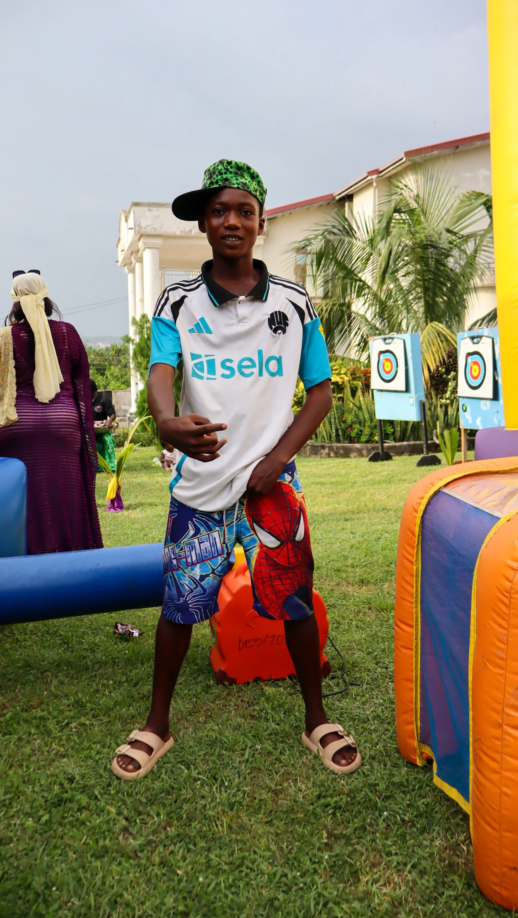 A young boy standing outdoors on a grassy area, wearing a sports jersey and shorts with a Spider-Man design, a green patterned cap, and beige sandals, with targets in the background.