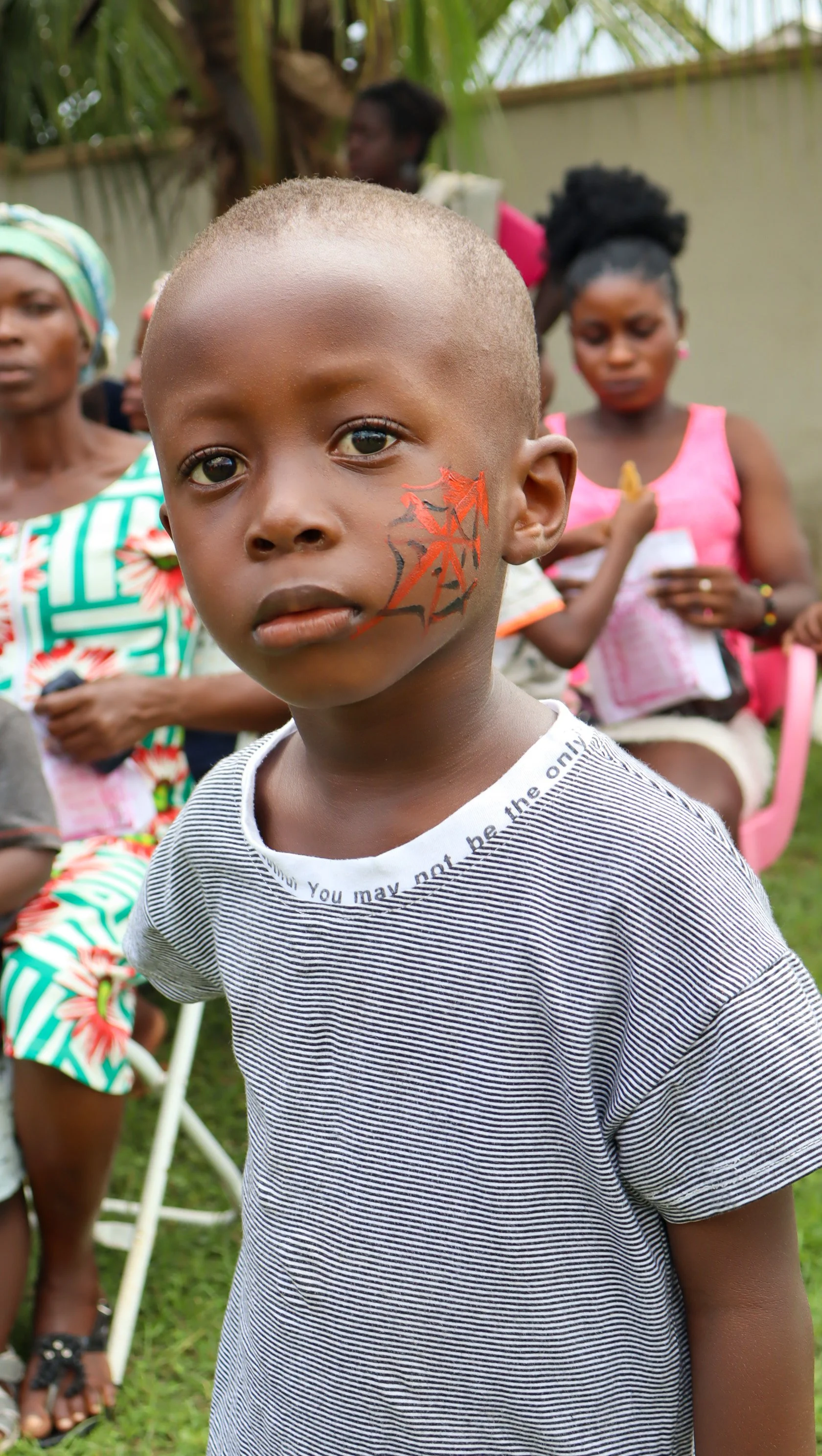 A young boy with a painted face standing outdoors surrounded by women, some wearing colorful clothing and headwraps, with a green leafy background.