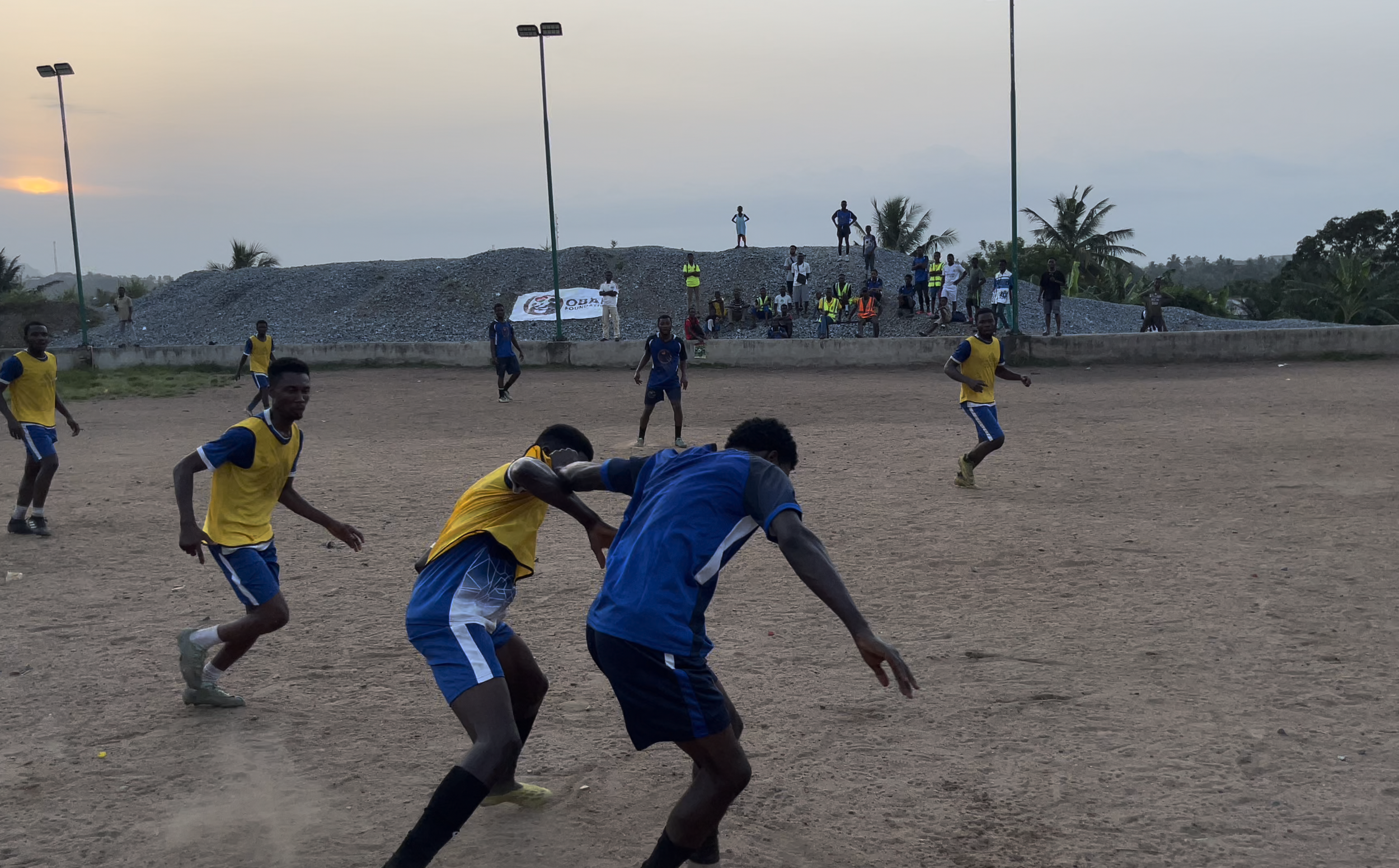 Soccer players in yellow and blue uniforms playing on a dirt field during sunset, with spectators and a rocky mound with palm trees in the background.