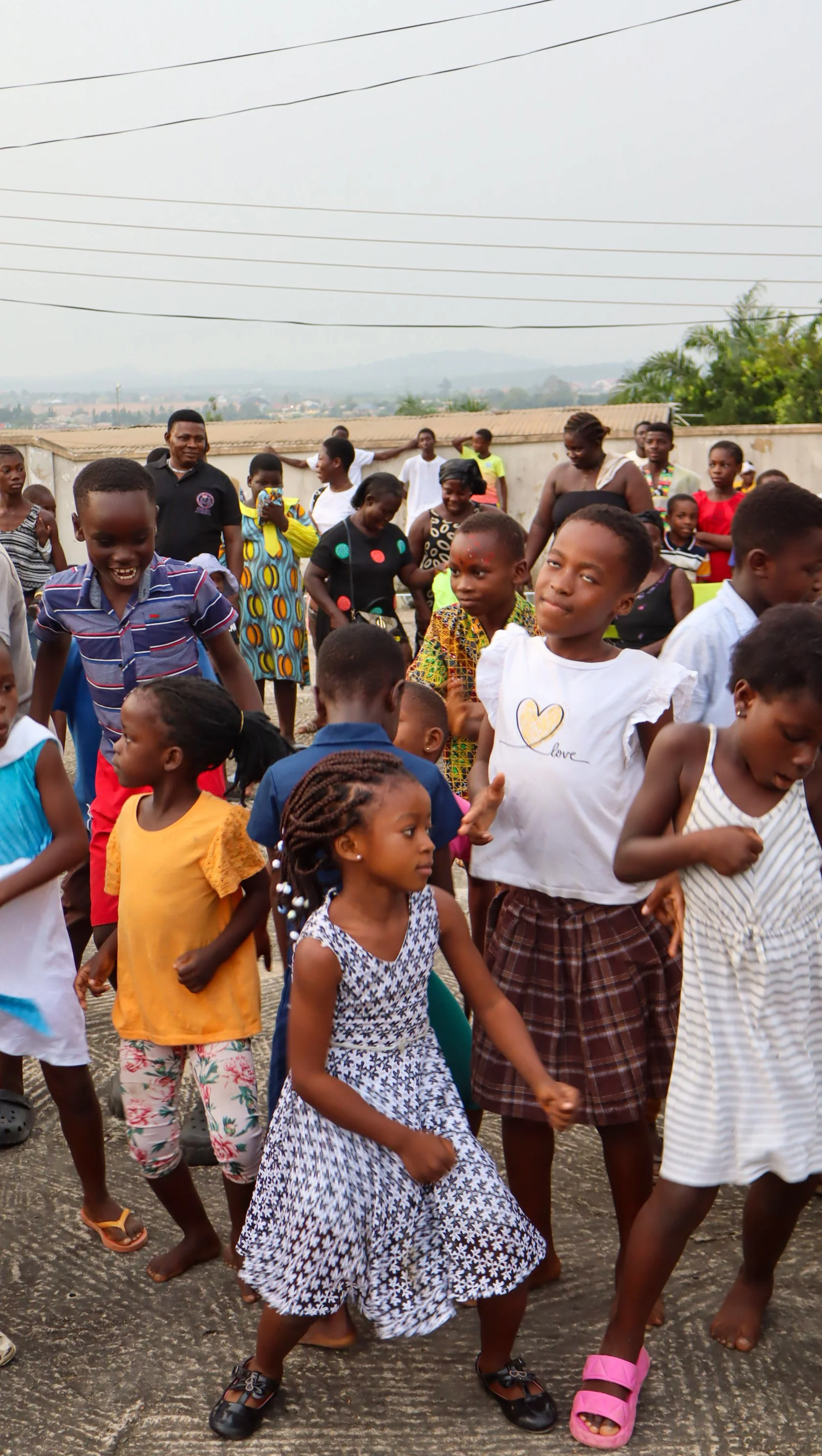 Children dancing outdoors, with adults watching in the background, on a cloudy day.