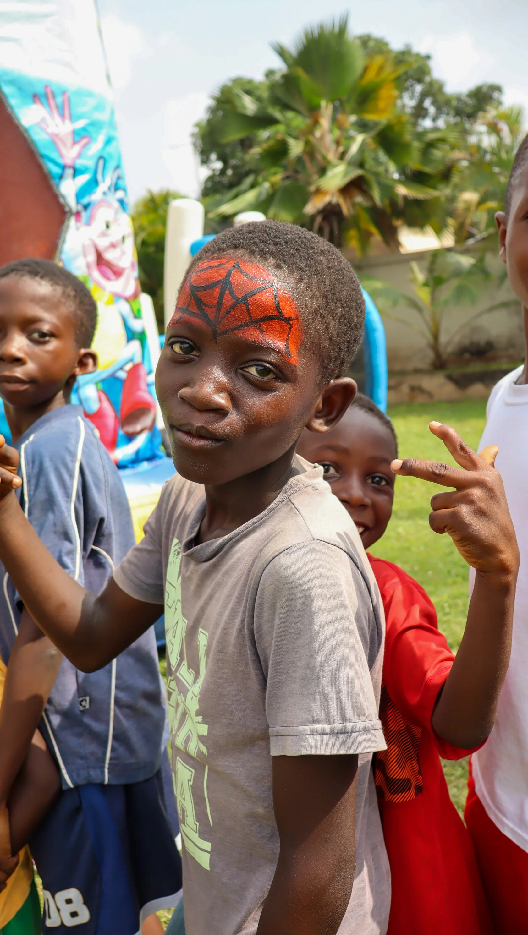 A group of children outdoors, with a boy in the center having face paint resembling a spider web on his forehead, in front of a colorful mural with cartoon characters and palm trees.