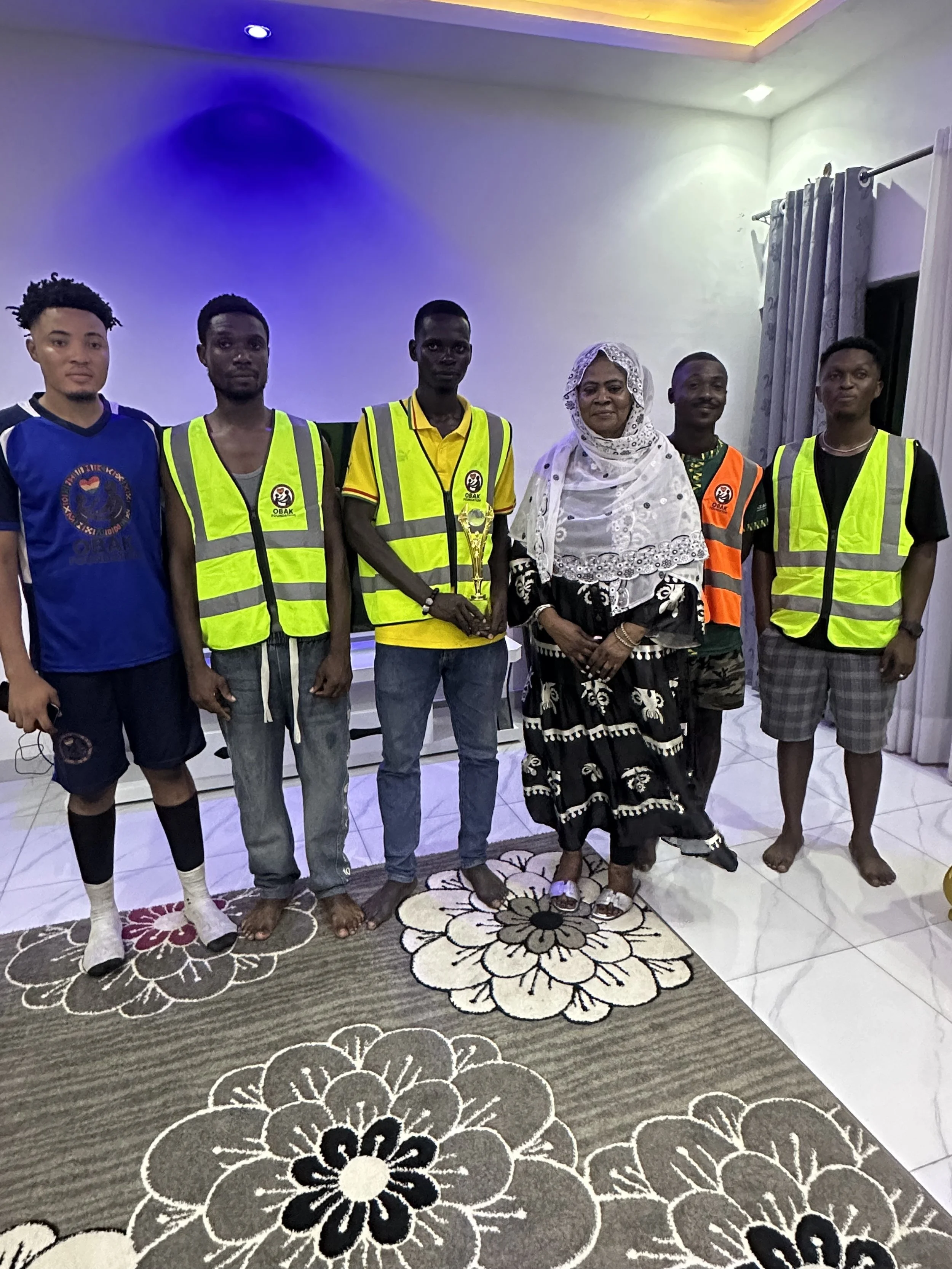 A group of six people standing indoors on a floral rug, including five young men wearing neon safety vests, one woman in a black and white dress with a lace headscarf, and one young man holding a small trophy, with a plain wall and curtains in the ba