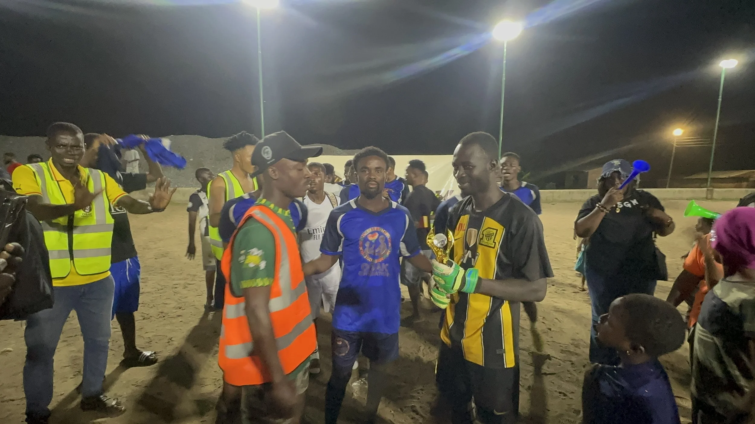 Group of soccer players and spectators gathered on a sandy field at night, celebrating. One player holds a trophy, while others applaud and cheer.