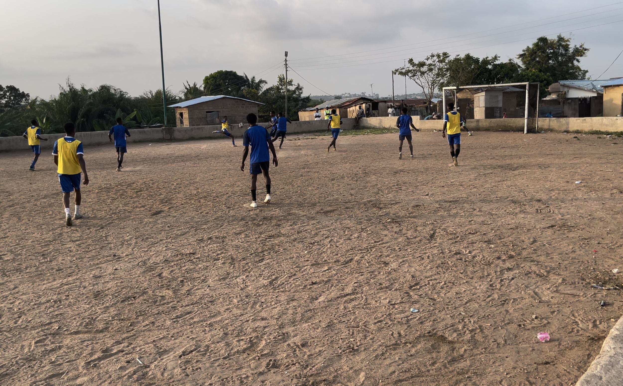 Children playing soccer on a dirt field in a village with small houses and trees in the background.