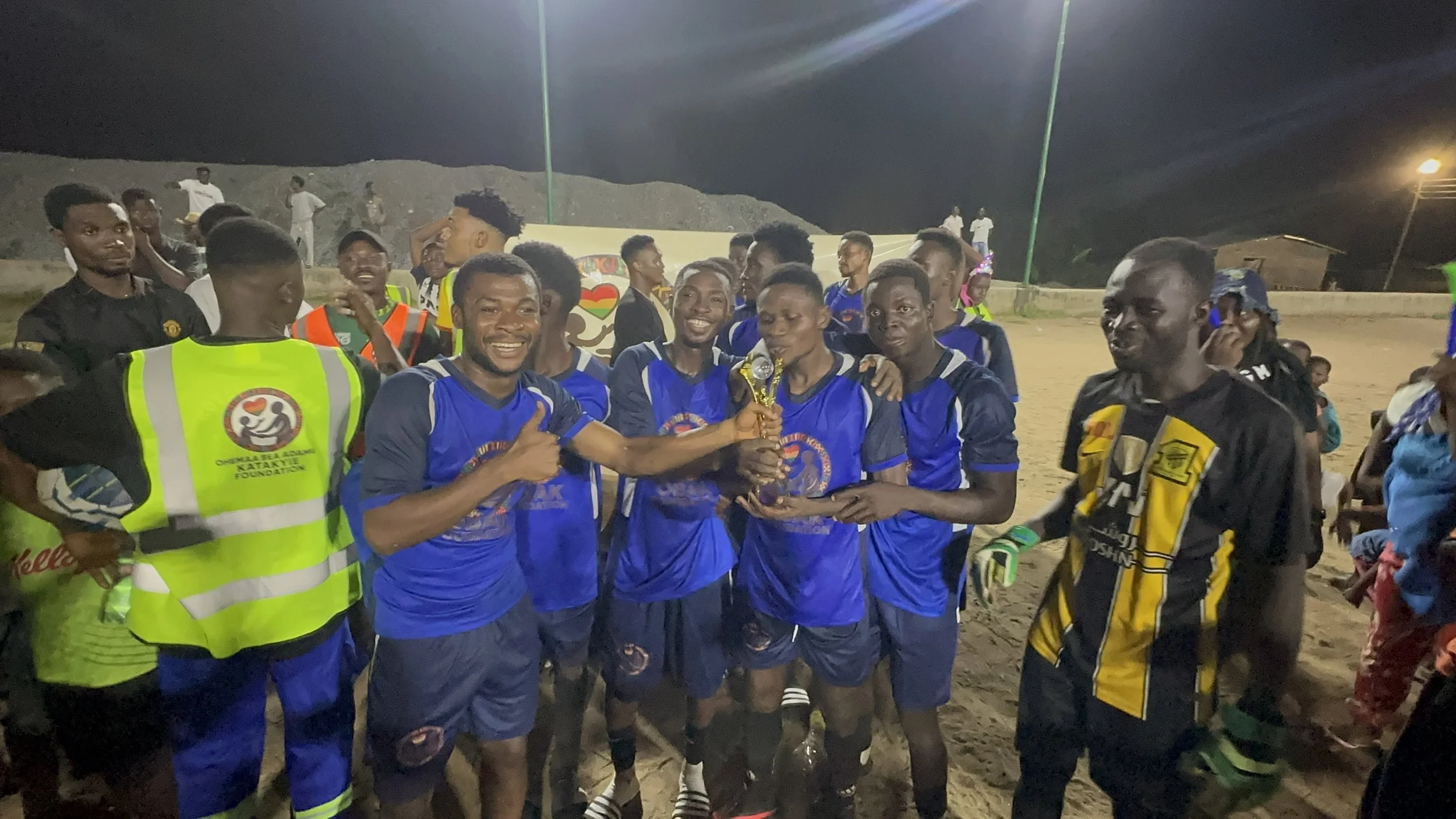 A group of young men in blue sports uniforms celebrate on a dirt field at night, holding a trophy, with smiling faces and some wearing jerseys with a logo. Others are in casual or safety vests, and there are additional people in the background.