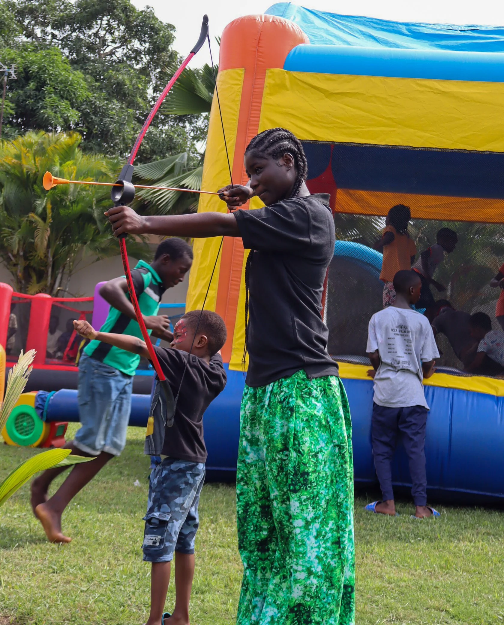 Children playing with a bow and arrow toy at a birthday party in a backyard with a bounce house in the background.
