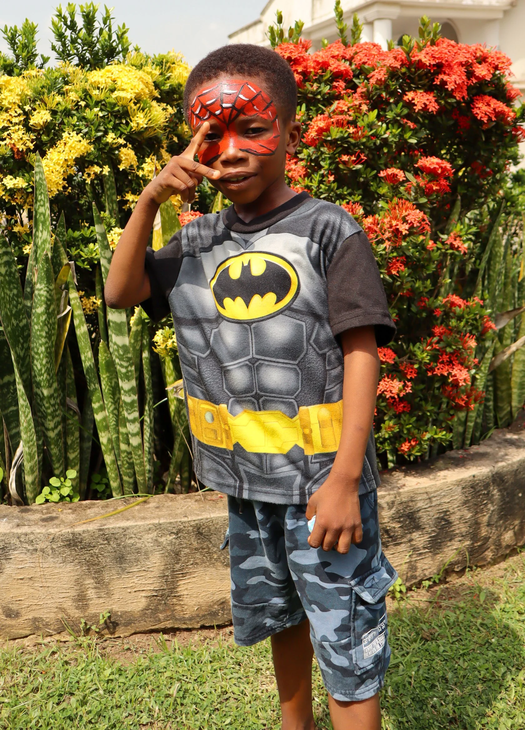 A young boy with face paint resembling Spider-Man, wearing a Batman-themed t-shirt and camouflage shorts, standing outdoors in front of colorful flowers, making a peace sign with his right hand.