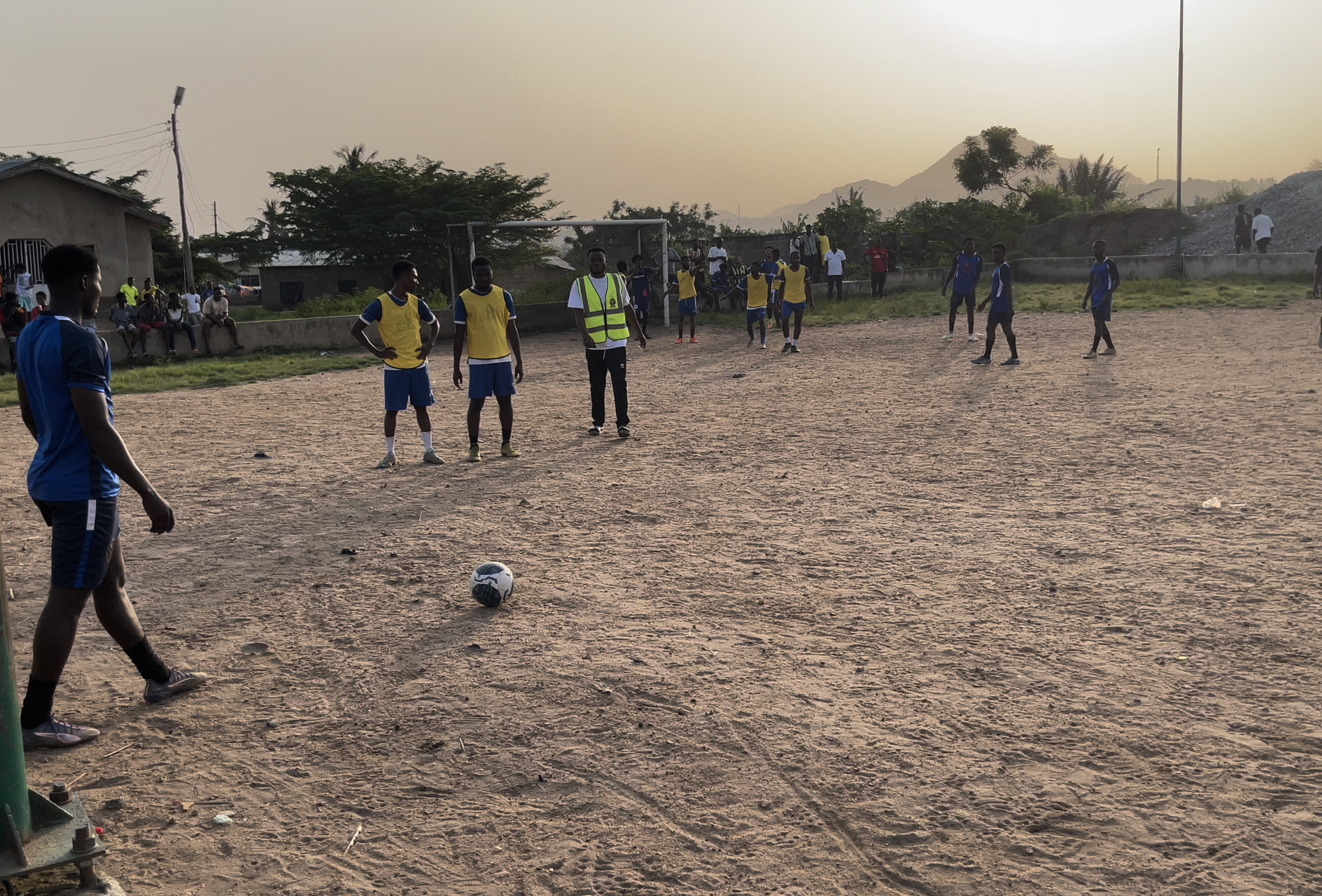 Young men playing soccer on a dirt field with spectators sitting on concrete walls and some standing in the background, with an outdoor setting and mountains in the distance during sunset.